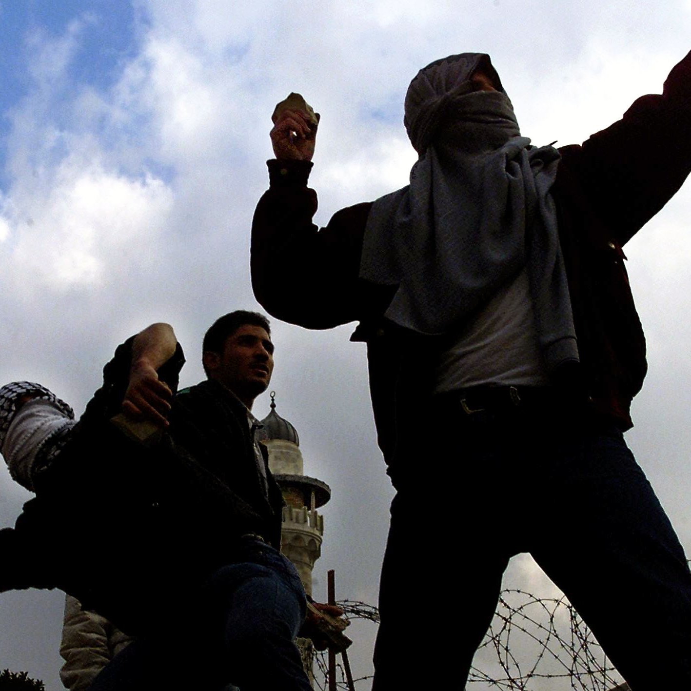 L'immagine mostra un gruppo di persone in una situazione di tensione, probabilmente in un contesto di protesta o conflitto. Alcuni individui sono coperti da sciarpe o fazzoletti, mentre sembrano lanciare dei proiettili, forse pietre. Sullo sfondo si possono vedere strutture architettoniche e un cielo nuvoloso, che contribuiscono all'atmosfera drammatica della scena. La presenza di filo spinato suggerisce una situazione di conflitto o di controllo militare.