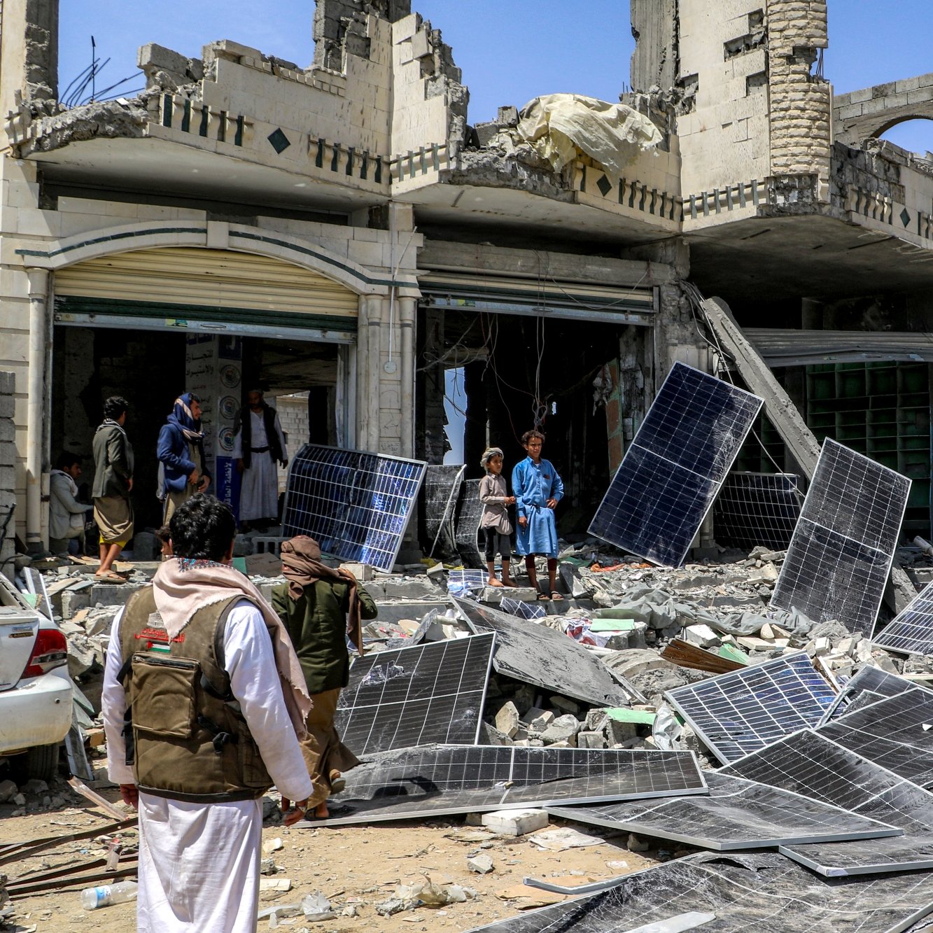 صورة لمكان مدمر، مباني مهدمة وألواح شمسية متكسرة، وأشخاص يتجمعون في الموقع.