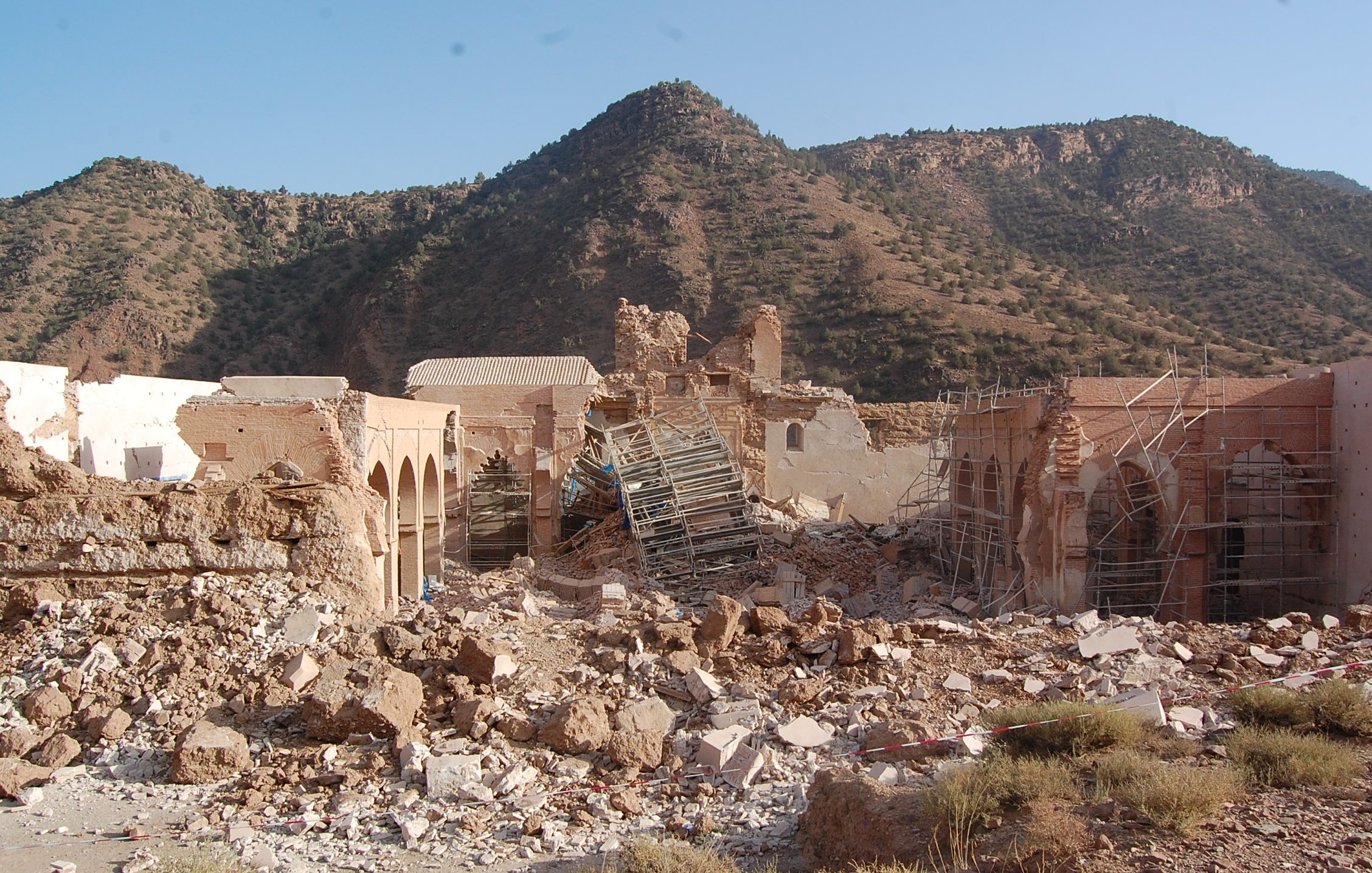 L'image montre un site de ruines où les structures sont partiellement effondrées, avec des débris éparpillés sur le sol. Des échafaudages sont visibles autour des bâtiments, suggérant un effort de restauration ou de démolition. En arrière-plan, on aperçoit une montagne aux pentes douces, recouverte de verdure, sous un ciel dégagé. L'ensemble dégage une impression de désolation mêlée à la beauté naturelle environnante.