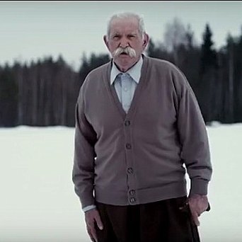 L'image montre un homme âgé se tenant debout sur un terrain enneigé. Il porte un pull en cardigan gris et un pantalon, et il a une moustache bien fournie. En arrière-plan, on peut voir une forêt de conifères qui ajoute une ambiance tranquille et froide à la scène. L'homme semble contemplatif, regardant droit devant lui dans un paysage hivernal.