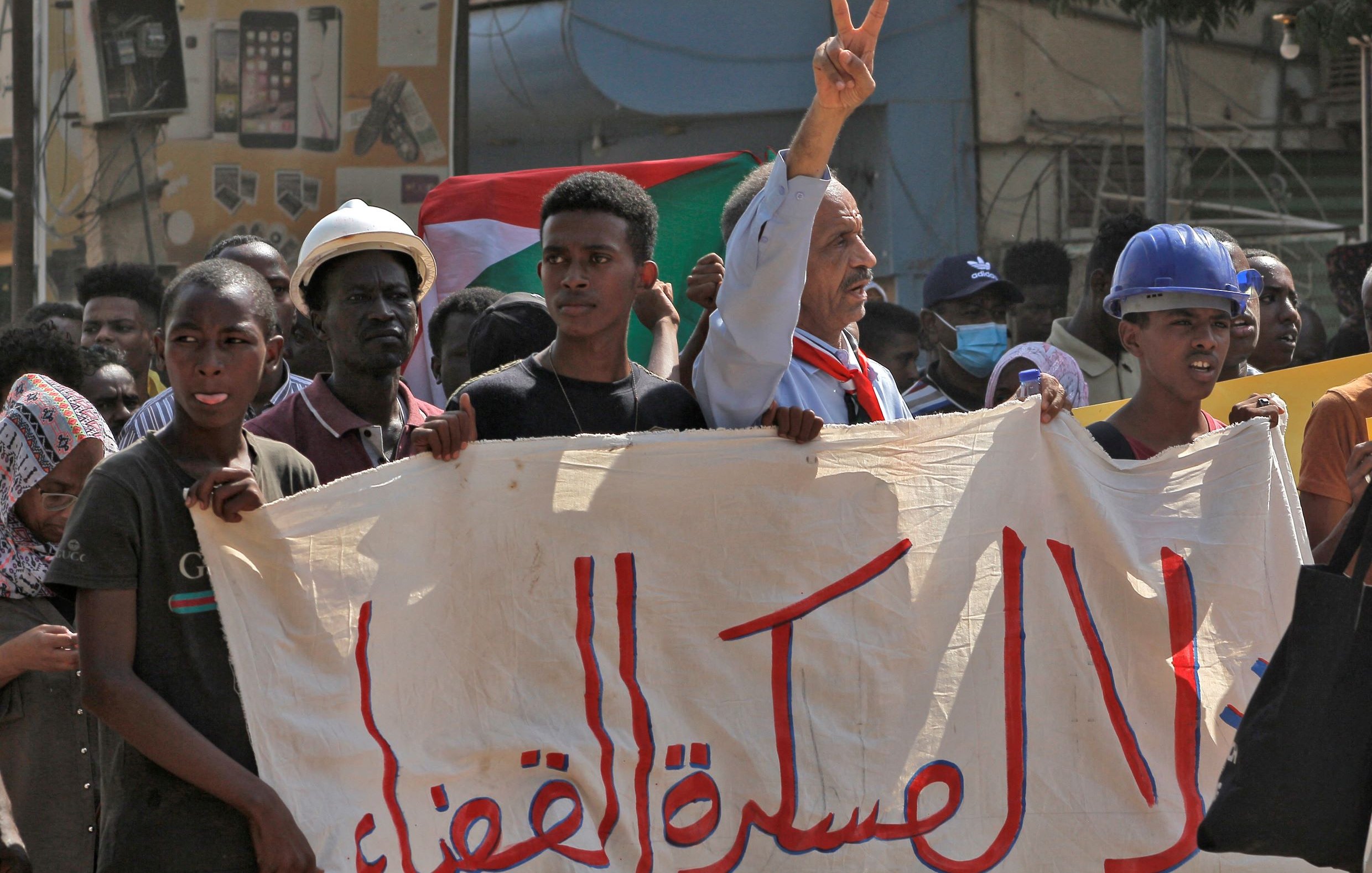 L'immagine mostra una manifestazione con diverse persone che sollevano cartelli e bandiere. In primo piano, un uomo con un cappello e una sciarpa al collo alza la mano mostrando il simbolo della pace. Gli altri manifestanti sembrano esprimere supporto per una causa, cominciando da una grande bandiera sudanese. Lo sfondo mostra dei cartelloni pubblicitari, mentre l'atmosfera appare carica di energia e determinazione.