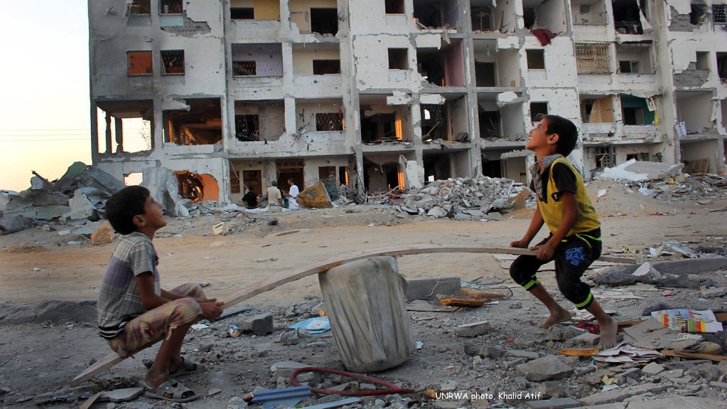 L'image montre deux enfants jouant à un jeu de bascule improvisé au milieu d'un paysage urbain en ruine. En arrière-plan, on peut voir un bâtiment endommagé, avec des fenêtres brisées et des murs effondrés, témoignant d'une destruction significative. Le sol est jonché de débris, et l'ambiance semble mélanger des éléments d'innocence enfantine et des conséquences tragiques d'un environnement en conflit. Les enfants semblent concentrés et engagés dans leur activité, apportant une note d'espoir malgré le contexte difficile.
