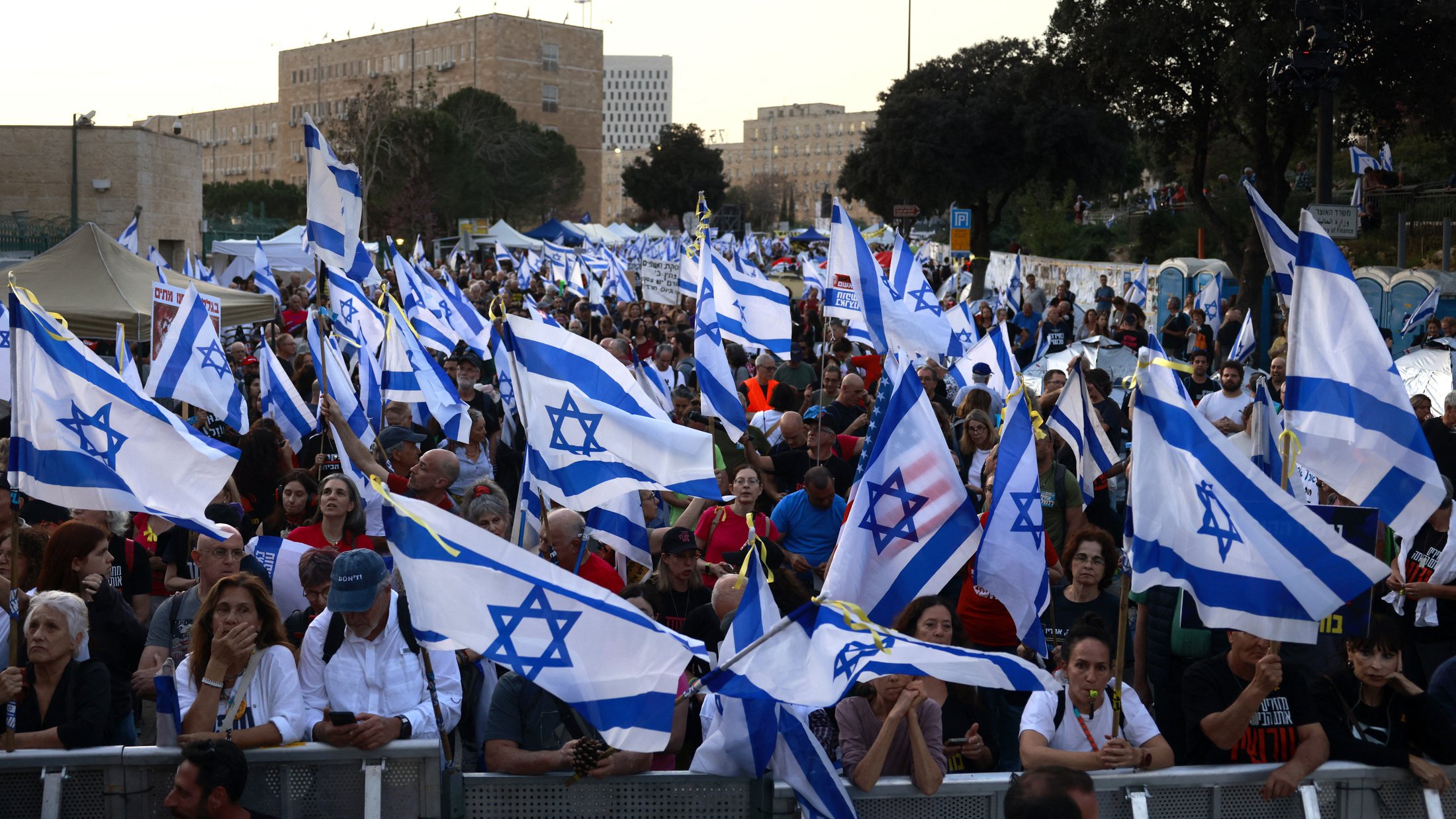 L'immagine mostra una folla di persone che partecipa a una manifestazione, sventolando molte bandiere di Israele. Il pubblico sembra essere molto numeroso e coinvolto, con alcuni partecipanti che usano telefoni cellulari. Sullo sfondo si possono vedere statue e edifici, mentre la scena è illuminata dalla luce del giorno. L'atmosfera appare vivace e carica di spirito collettivo.