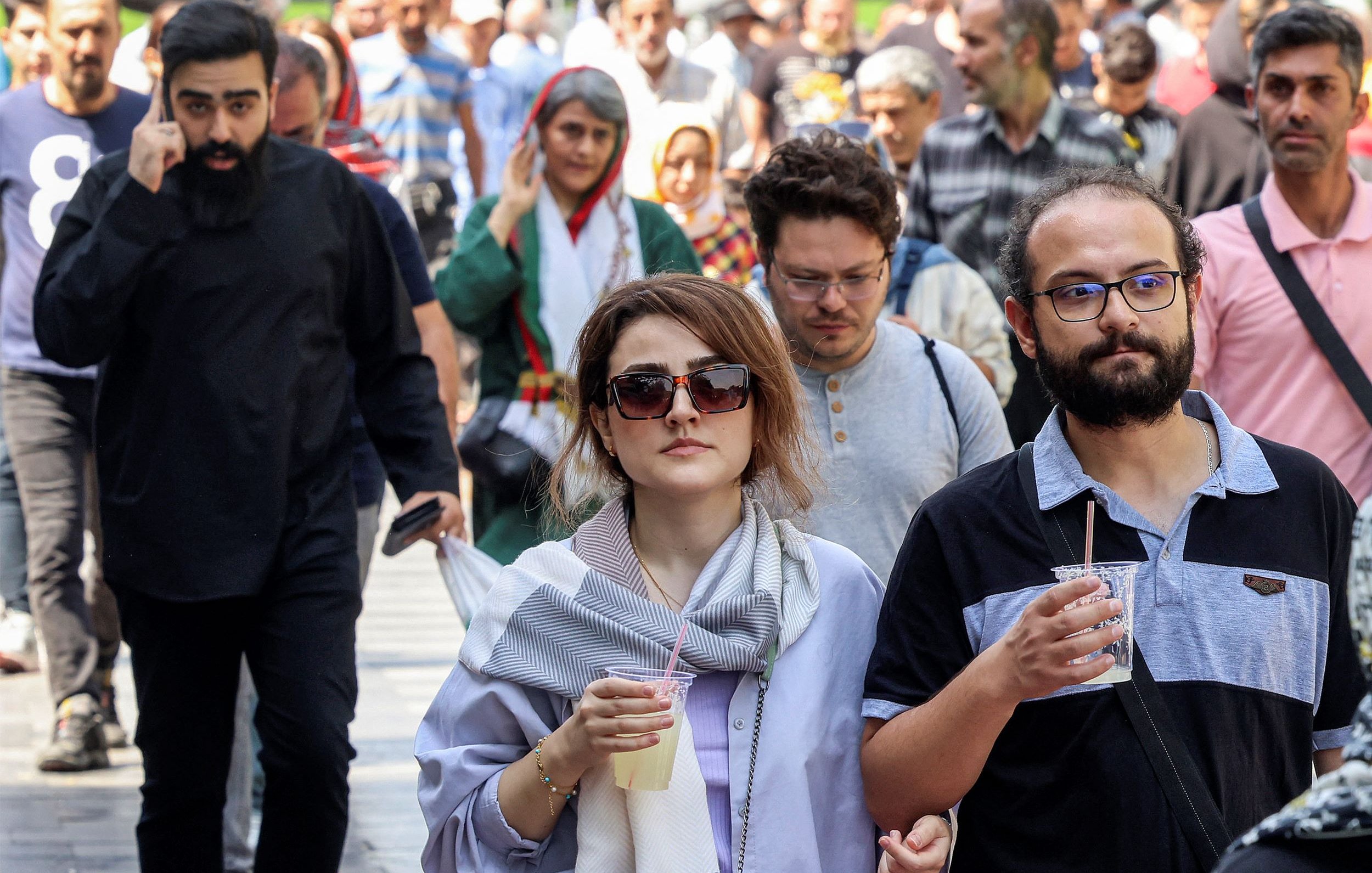 L'image montre une scène animée d'une rue où plusieurs personnes marchent. Au premier plan, deux personnes se déplacent côte à côte. Un homme portant des lunettes et une chemise à rayures tient un verre. À côté de lui, une femme avec des lunettes de soleil et une écharpe semble s'engager dans une conversation. En arrière-plan, d'autres personnes marchent, créant une atmosphère dynamique. On peut voir une variété de styles vestimentaires, suggérant une diversité culturelle.