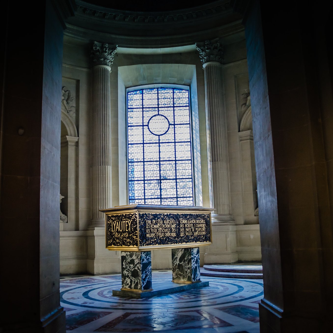 L'image montre une pièce intérieure d'un bâtiment majestueux, probablement un palais ou un musée. Au centre, il y a un grand meuble ou une caisse ornée de motifs décoratifs dorés et peints. Les murs sont en pierre, avec de très grandes fenêtres, à travers lesquelles entre une lumière douce, créant une atmosphère solennelle. Les colonnes et le sol richement décoré ajoutent à la grandeur de l'espace.