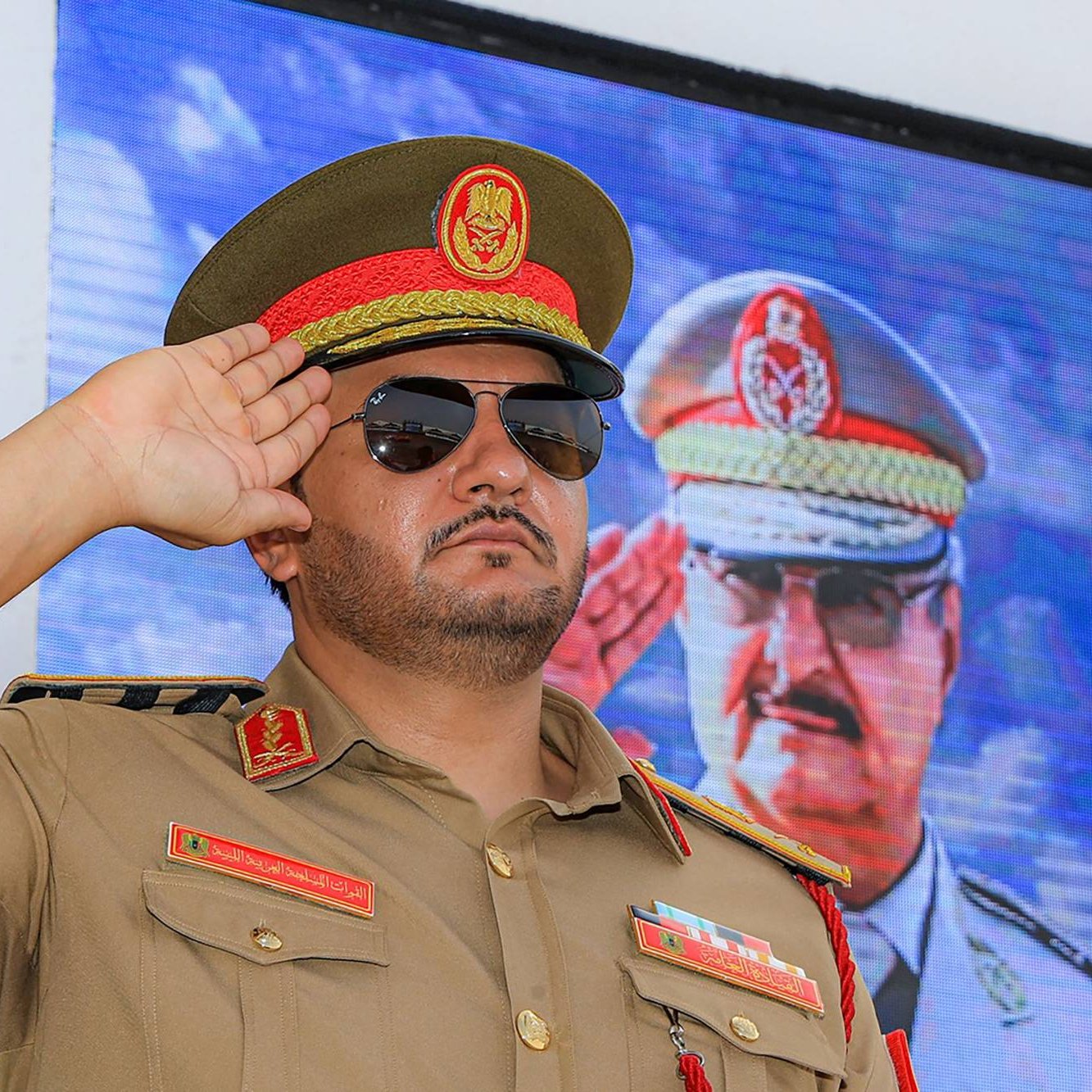 Un homme en uniforme militaire, lunettes noires, salue devant un écran.
