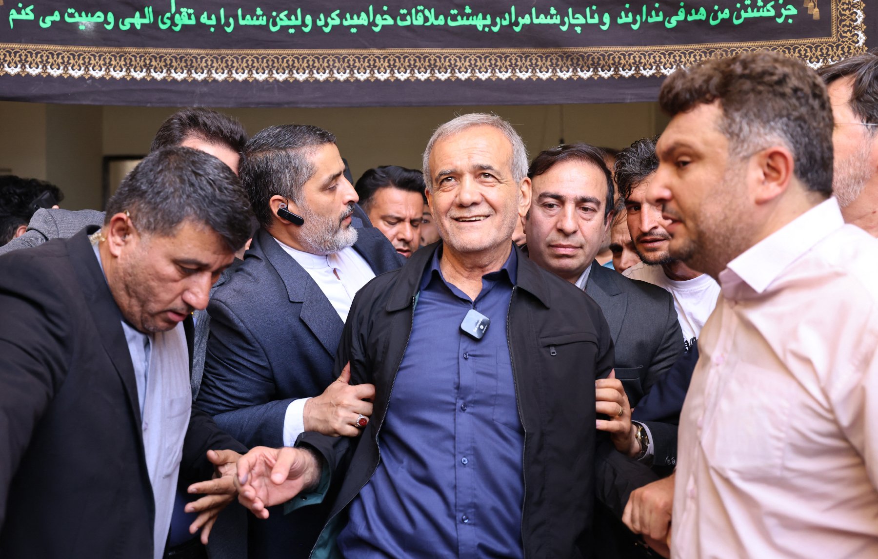 در این تصویر، گروهی از مردان در یک محیط اجتماعی دور یک مرد میانسال جمع شده‌اند. این مرد لبخند بر لب دارد و به نظر می‌رسد در حال برقراری ارتباط با دیگران است. محیط تصویر نشان‌دهنده‌ی یک رویداد مهم یا تجمع است. مردان اطراف او به نظر می‌رسند که به او احترام گذاشته و از او حمایت می‌کنند. متن در بالای تصویر به زبان فارسی نوشته شده است و به نظر می‌رسد حاوی یک پیام یا عنوان خاص باشد.