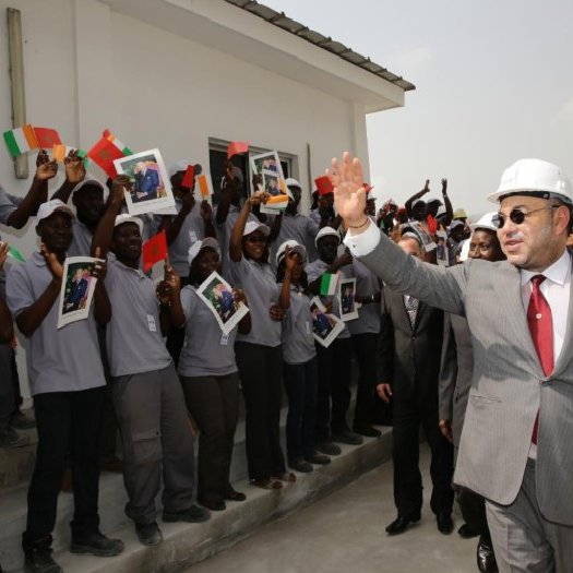 L'image montre un groupe de personnes, probablement des travailleurs ou des étudiants, levant des affiches avec des photos tout en saluant une personne qui semble être un dirigeant. Ce dernier porte un casque de sécurité et coûte en costume, indiquant une visite d'inspection ou une inauguration. Les personnes en arrière-plan sont toutes habillées de manière uniforme et semblent enthousiastes, créant une atmosphère de célébration. L'environnement est extérieur, avec un bâtiment en arrière-plan.