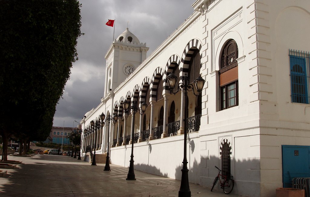 L'image montre un bâtiment architectural imposant, probablement un hôtel de ville ou un bâtiment administratif, avec une façade blanche. Ce bâtiment présente des arches décoratives et des fenêtres ornées. On peut apercevoir une grande horloge sur la tour, ainsi qu'un drapeau turc flottant au sommet. Le ciel est nuageux, ajoutant une ambiance dramatique à la scène. Des lampadaires sont alignés le long de la rue pavée qui borde le bâtiment, et un vélo est garé sur le côté. Des arbres bien taillés sont également visibles, contribuant à l'esthétique générale de l'endroit.