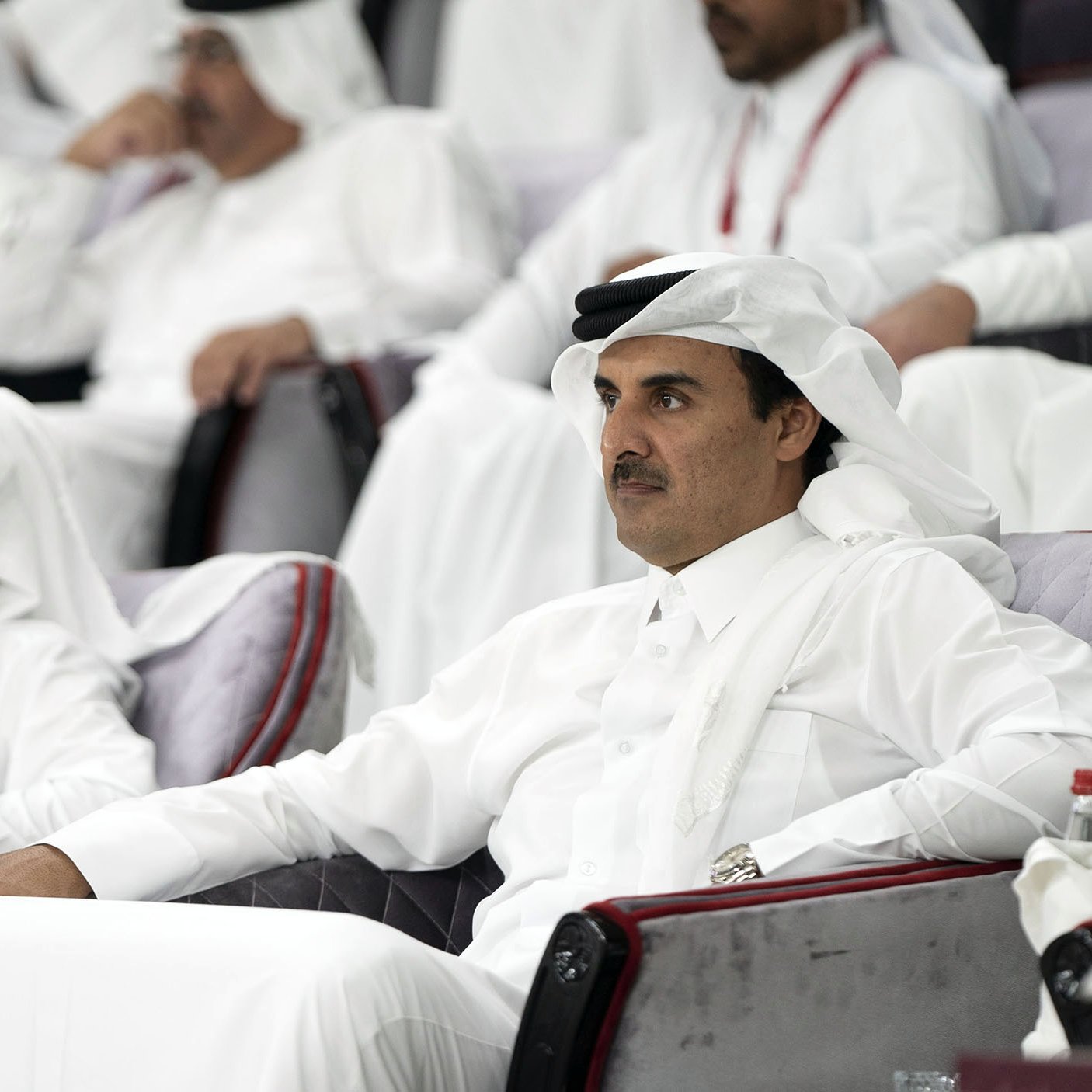 L'image montre un groupe de personnes assises dans des sièges, probablement dans un stade ou un lieu événementiel. Deux hommes sont au premier plan, vêtus de vêtements traditionnels qataris, avec des turbans blancs et des robes longues. Ils semblent attentifs et engagés dans l'événement se déroulant devant eux. L'atmosphère est celle d'un rassemblement formel, avec d'autres personnes en arrière-plan également habillées de manière traditionnelle.