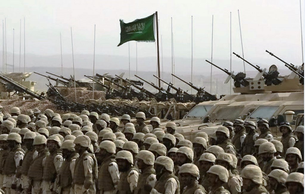 تظهر في الصورة مجموعة من الجنود يرتدون زيًا عسكريًا واقفين في صفوف منتظمة. خلفهم، توجد دبابات ومدرعات عسكرية، حيث تُشاهد المدافع موجهة نحو الأمام. في خلفية الصورة، يوجد علم أخضر يرفرف، مما يوحي بأن هذه الصورة مرتبطة بقوة عسكرية أو استعراض. تعكس الصورة أجواءً عسكرية وهيبة القوات المدافعة.