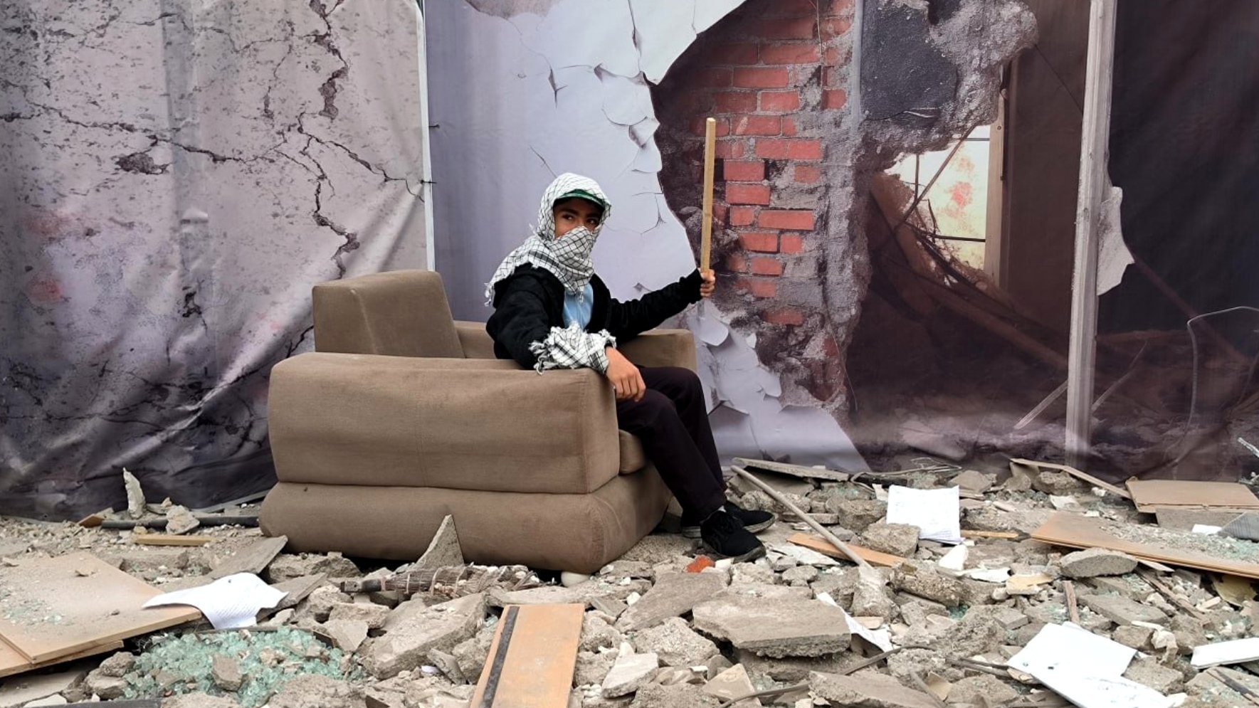 L'image montre une scène de destruction avec des débris éparpillés au sol, tels que des morceaux de béton et des morceaux de verre brisé. Au centre, une personne est assise sur un canapé apparemment plus ou moins intact. Elle porte un foulard autour de la tête et tient un bâton. En arrière-plan, il y a une illustration d'un mur brisé, ajoutant à l'impression de désolation et de chaos de la scène.