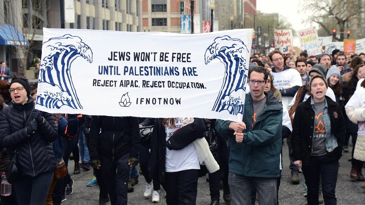 The image shows a group of protesters marching together, holding a large banner that reads, "Jews won't be free until Palestinians are. Reject AIPAC. Reject occupation." The banner features artistic waves, and the participants appear to be actively engaged in the demonstration, dressed in casual winter clothing. The background includes urban buildings and other protesters. The atmosphere conveys a sense of solidarity and activism focused on social justice issues.