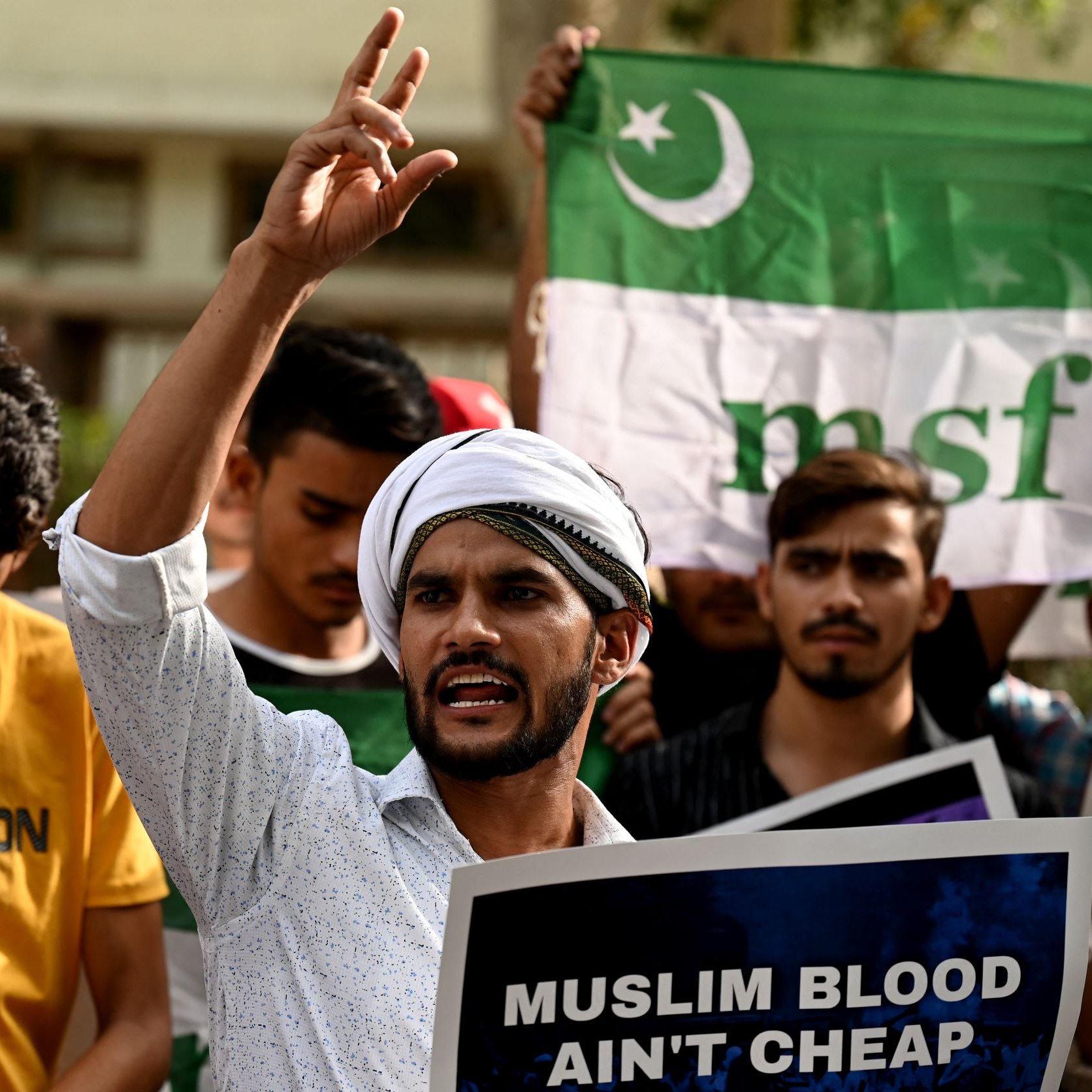 L'image montre un groupe de manifestants rassemblés, tenant des pancartes et des drapeaux. Au premier plan, un homme s'exprime avec passion, levant un doigt en l'air. Il porte une chemise claire et un turban. Les autres participants dans le fond semblent également engagés dans la manifestation, brandissant des affiches avec des messages. L'atmosphère semble intense et revendicative.