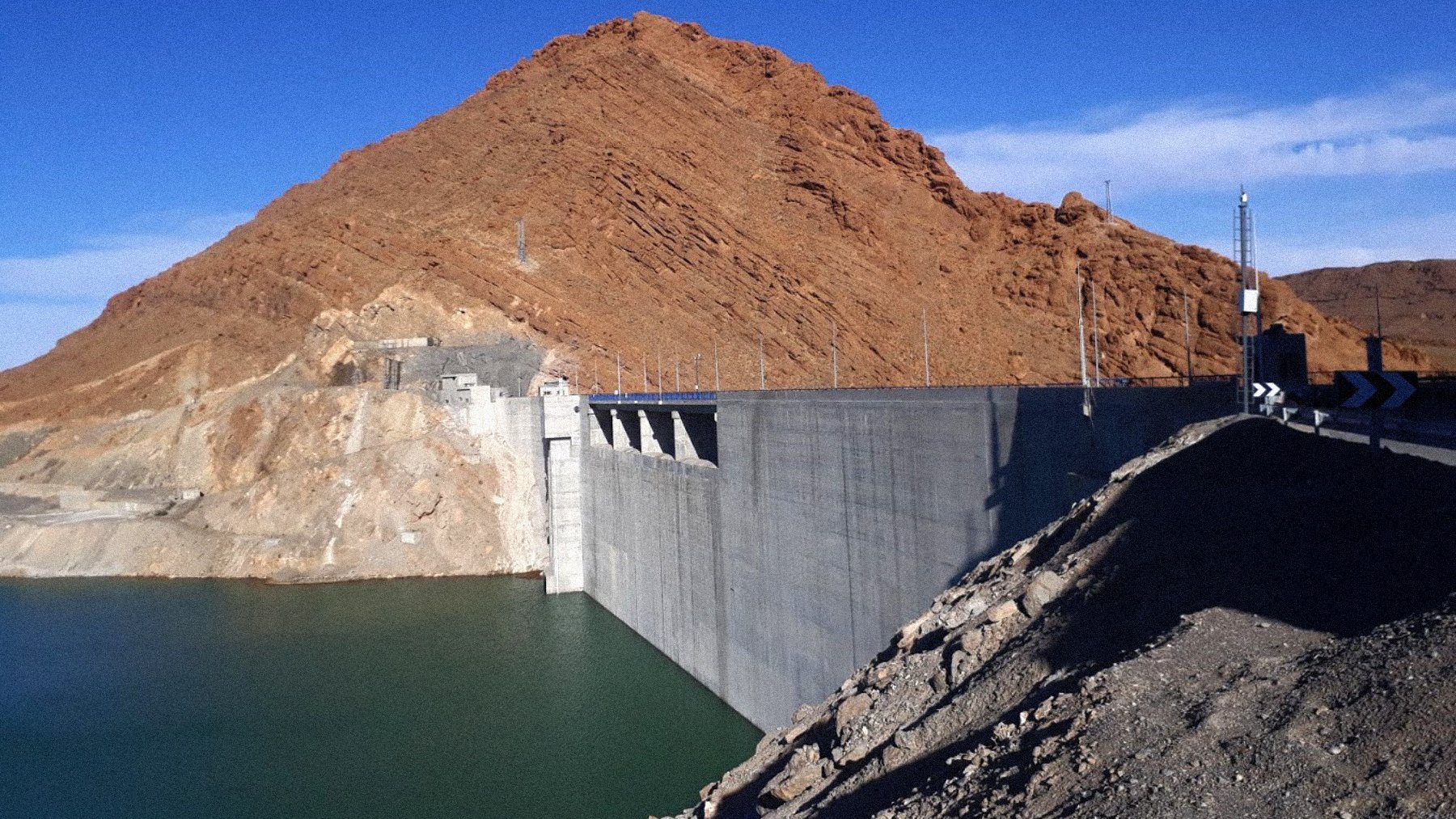 L'image montre un barrage construit sur un cours d'eau, avec une grande structure en béton qui s'étend le long de la rive. À l'arrière-plan, on voit une montagne aride, aux pentes abruptes et aux teintes brunes. Le ciel est dégagé, d'un bleu vif, et l'eau du réservoir est calme et d'une teinte verte-bleue. Le paysage semble sec et stérile, typique des régions désertiques.