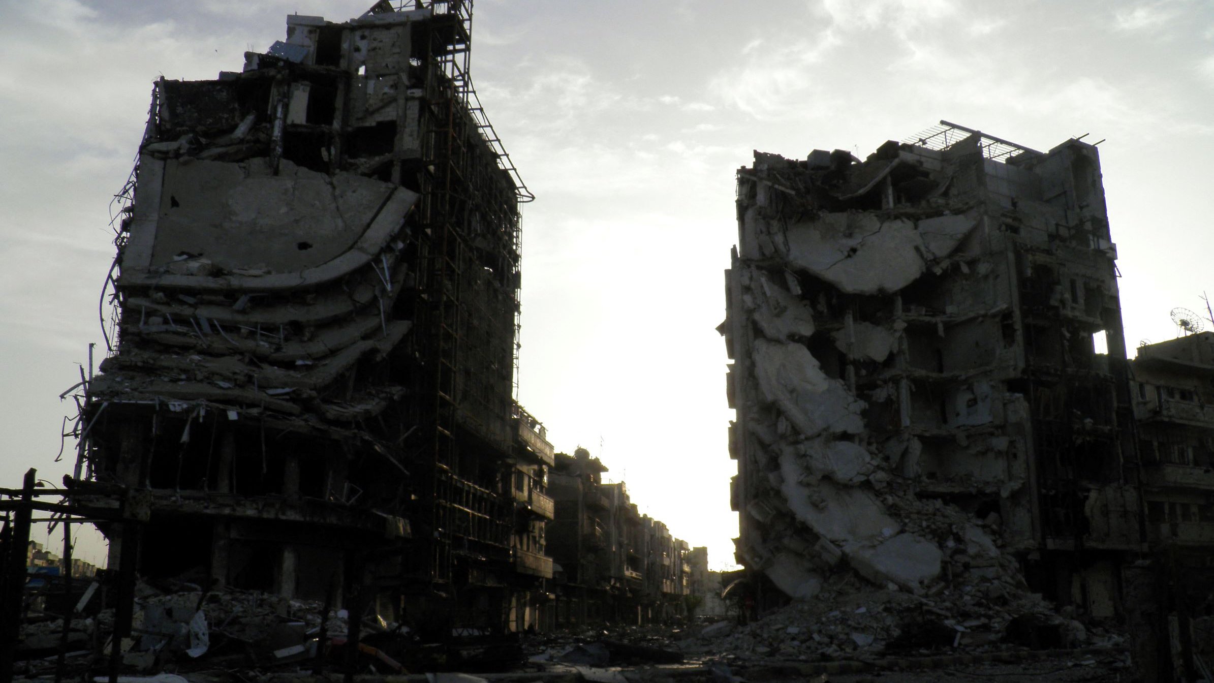 تظهر الصورة عمارتين محطمتين في منطقة يبدو أنها تعرضت لتدمير واسع. المباني مشوهة، حيث تظهر الجدران المنهارة والأسطح المدمرة، مما يعكس آثار الدمار. السماء في الخلفية تبدو ملبدة، مما يضيف شعورًا بالخيبة والخراب. المنطقة المحيطة يبدو أنها مهجورة، مما يعكس الأثر العميق للصراعات أو الكوارث التي أدت إلى هذا الوضع.