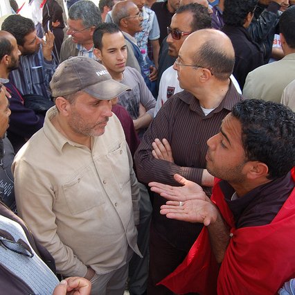 L'image montre un groupe de personnes rassemblées dans un endroit public, probablement lors d'une manifestation ou d'un rassemblement. Plusieurs hommes se tiennent ou se parlent les uns aux autres, affichant des expressions variées qui semblent indiquer une discussion animée. Certains portent des vêtements décontractés et il y a une ambiance de dialogue, avec des gestuelles qui suggèrent l'échange d'opinions. En arrière-plan, on peut apercevoir d'autres personnes qui regardent la scène. L'atmosphère semble engageante et dynamique.