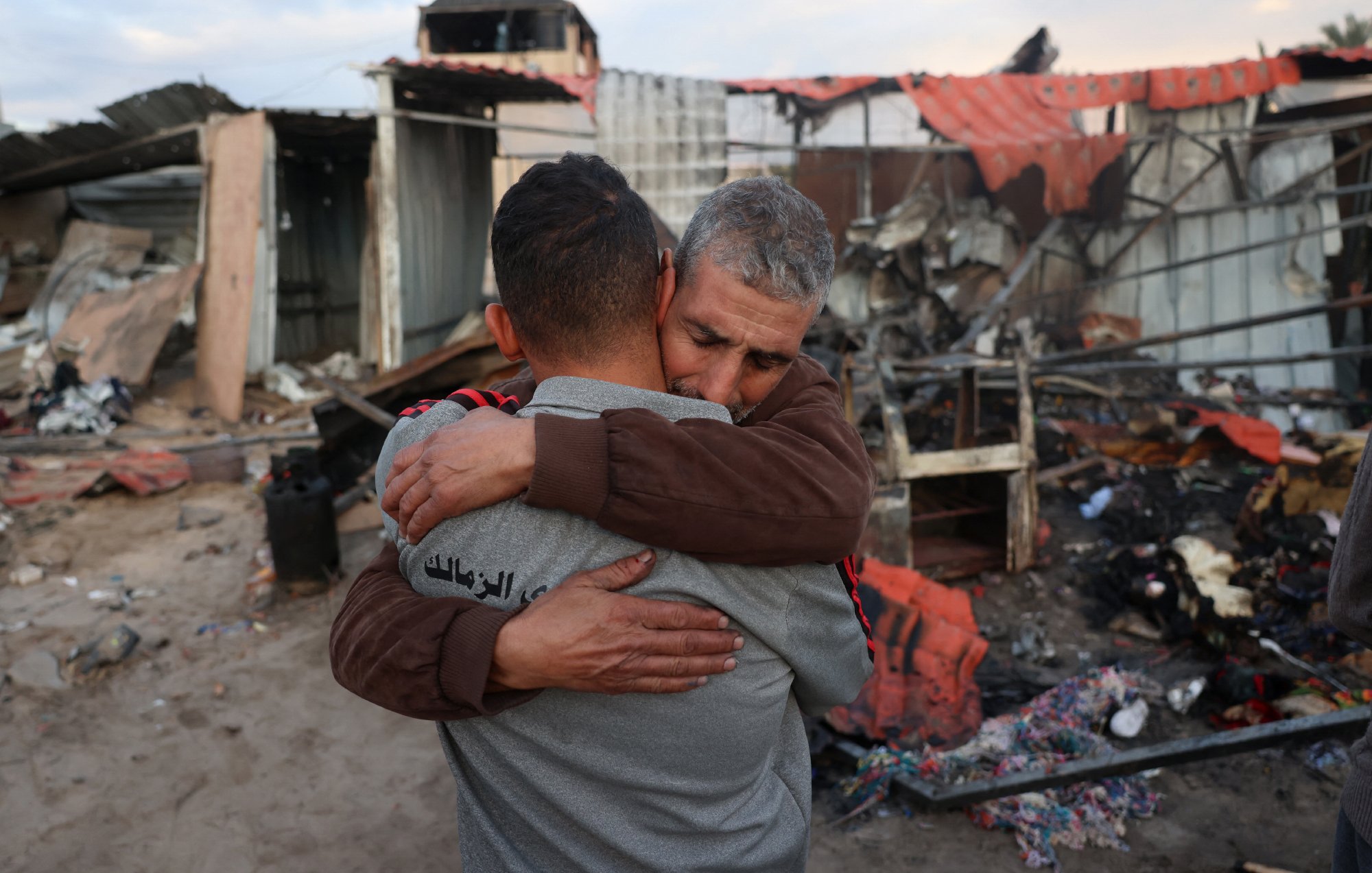 L'immagine mostra due uomini che si abbracciano in un contesto distrutto. Sullo sfondo, si possono vedere i resti di edifici danneggiati, con detriti e rovine sparse. La scena trasmette un forte senso di emozione e solidarietà tra le due persone, che si sostengono in un momento difficile. La luce naturale del cielo aggiunge un'atmosfera malinconica alla situazione.