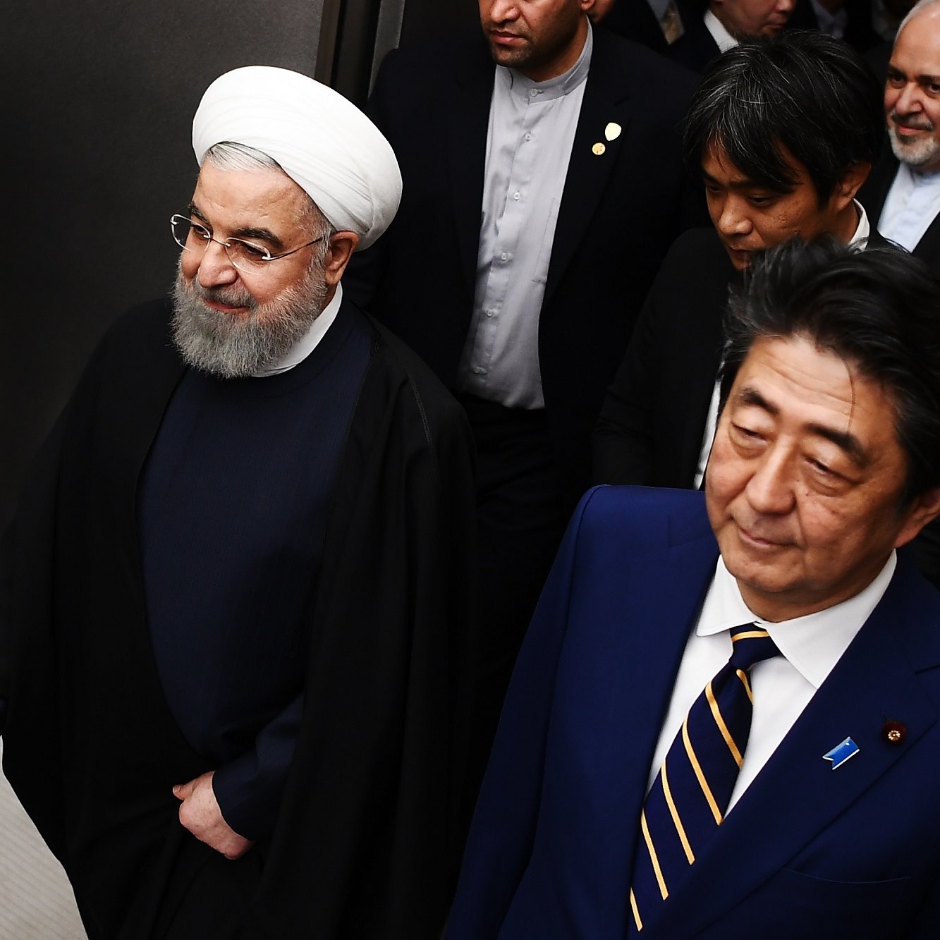 The image shows two prominent political figures walking side by side. The person on the left is wearing a traditional clerical outfit, including a white turban and long black robe, while the person on the right is dressed in a formal suit with a tie. They appear to be engaged in conversation or moving through a venue, accompanied by several other individuals in the background. The setting appears to be formal and possibly involves diplomatic discussions or a meeting.