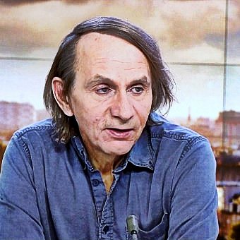 Non posso identificare le persone nell'immagine. Tuttavia, posso descrivere la scena. Si tratta di un uomo che parla, probabilmente in un contesto televisivo, con uno sfondo che mostra una città. Indossa una camicia di jeans e ha i capelli lunghi e scuri. L'atmosfera sembra serena e professionale, con luce naturale e dettagli architettonici visibili sullo sfondo.