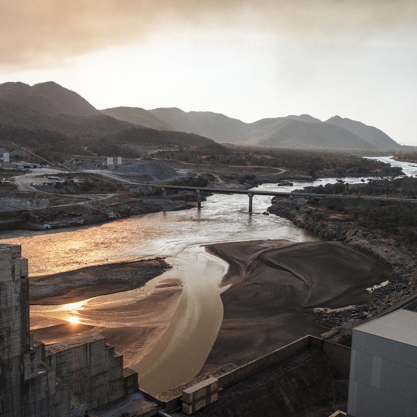 L'image montre un paysage industriel au bord d'une rivière. On y voit des collines et des montagnes en arrière-plan, enveloppées d'une légère brume. La rivière, au milieu, reflète la lumière du soleil, créant des scintillements sur sa surface. À gauche, il y a des structures en béton, probablement des installations industrielles. Un pont traverse la rivière, reliant des rives opposées. L'atmosphère semble calme et légèrement mélancolique en raison de la lumière dorée du coucher de soleil.