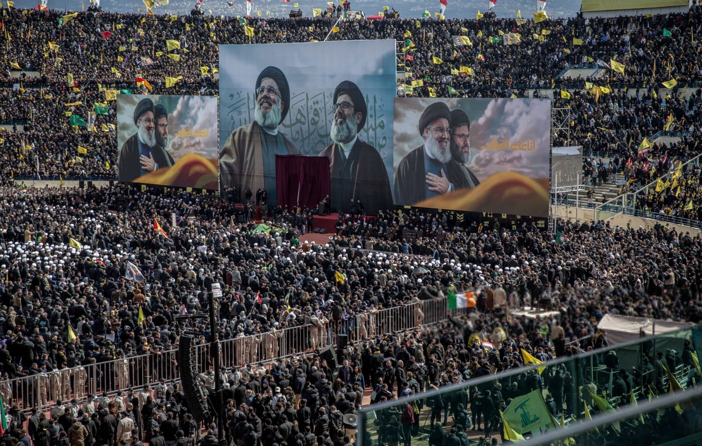 L'image montre une grande foule rassemblée dans un stade, avec des étendues de drapeaux de différentes couleurs. Au centre, trois grandes affiches de figures importantes sont visibles, probablement des leaders religieux ou politiques. L'atmosphère semble festive et engagée, avec des personnes portant des chapeaux ou des foulards spécifiques, suggérant une démonstration de soutien ou de solidarité. Des bannières et des drapeaux supplémentaires peuvent également être aperçus, accentuant l'aspect coloré et vibrant de l'événement.
