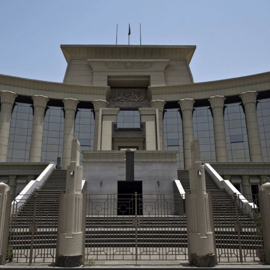 L'image montre un bâtiment imposant, probablement une institution judiciaire, avec une architecture moderne. La façade est ornée de colonnes majestueuses et de grandes fenêtres réfléchissantes, créant une impression de grandeur. Il y a des escaliers menant à l'entrée principale, qui semble être encadrée par des éléments décoratifs. Le ciel est dégagé, suggérant une journée ensoleillée. Des palmiers sont visibles sur les côtés, ajoutant une touche tropicale à l'environnement.