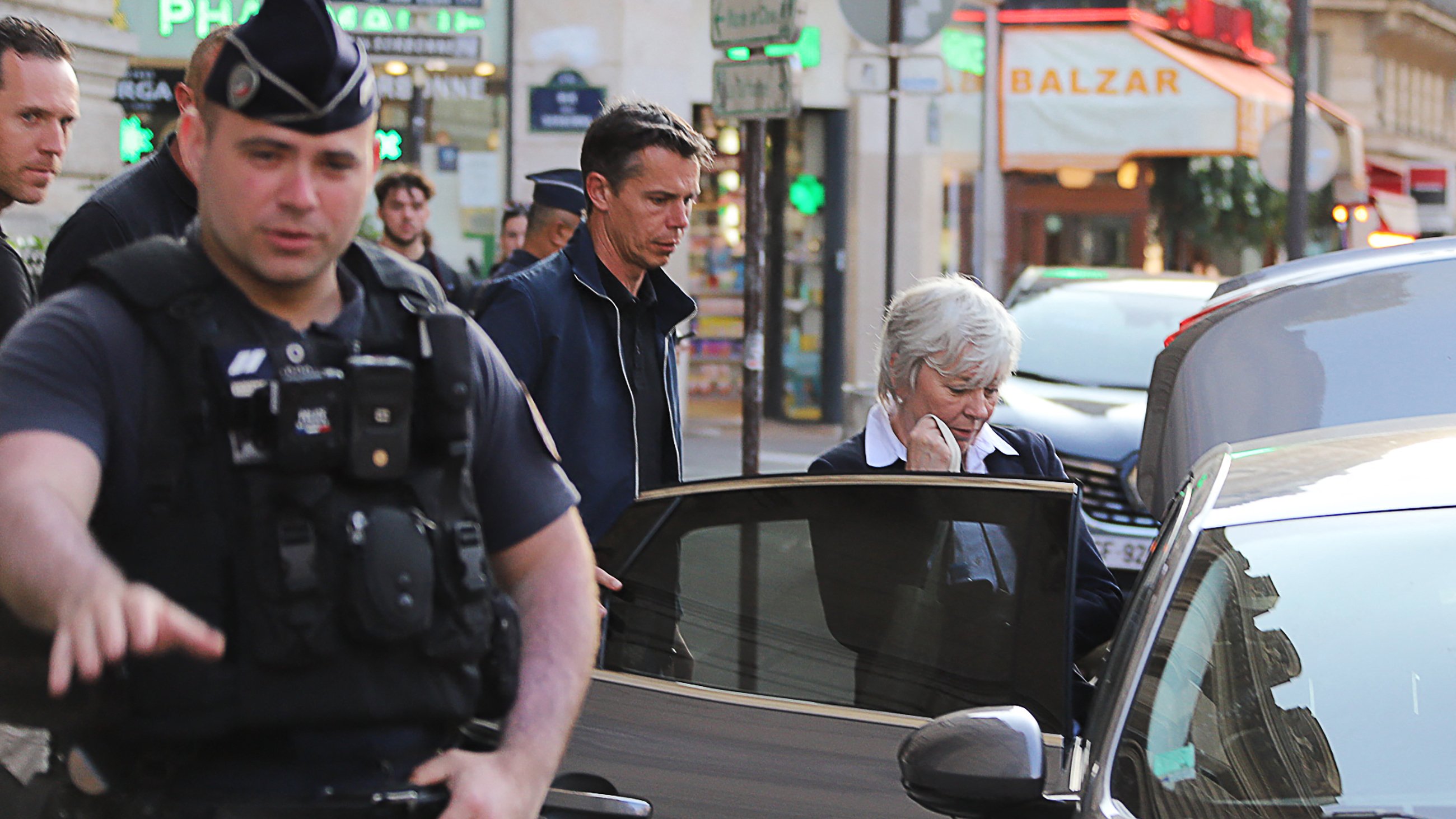 The image shows a scene on a city street. In the foreground, a police officer in uniform appears to be directing traffic or overseeing an incident. To the right, there are two individuals: a man in a dark jacket and a woman with short white hair, who seems to be getting into a car. The background features buildings and some signage, suggesting a busy urban environment. The atmosphere looks quite active, likely indicative of a recent event or situation requiring police presence.
