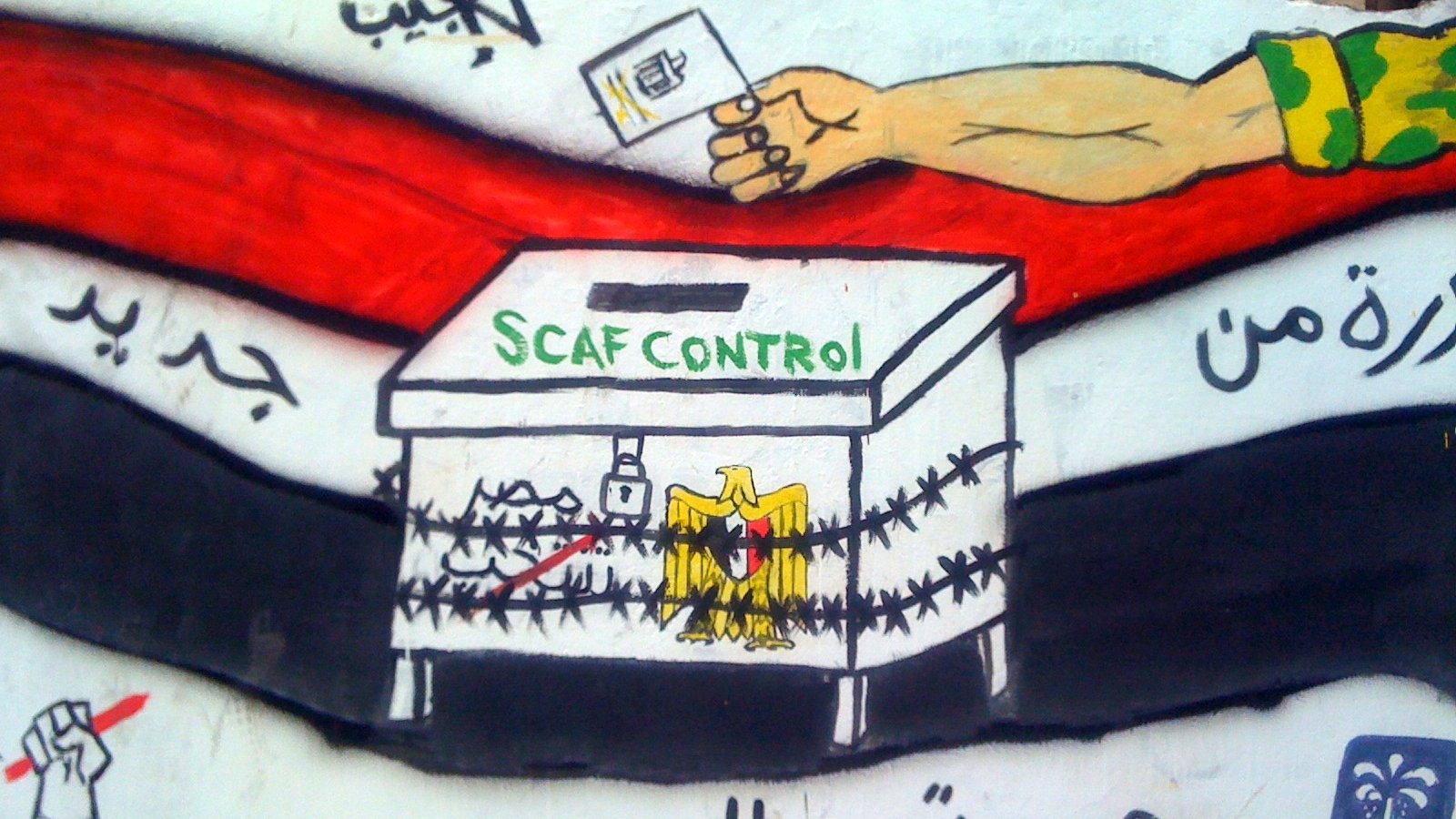 L'image montre une fresque murale avec une boîte de vote entourée de barbelés. Au-dessus de la boîte, on peut voir la phrase "SCAF CONTROL" écrite en vert. À gauche, il y a un poing levé, symbolisant la résistance ou la lutte, et à droite, on peut apercevoir des symboles qui représentent probablement des idéaux de liberté et de changement. La fresque utilise des couleurs vives comme le noir, le rouge et le blanc, qui sont souvent associés à des mouvements politiques. Le message semble critiquer ou commenter le contrôle militaire sur le processus électoral.