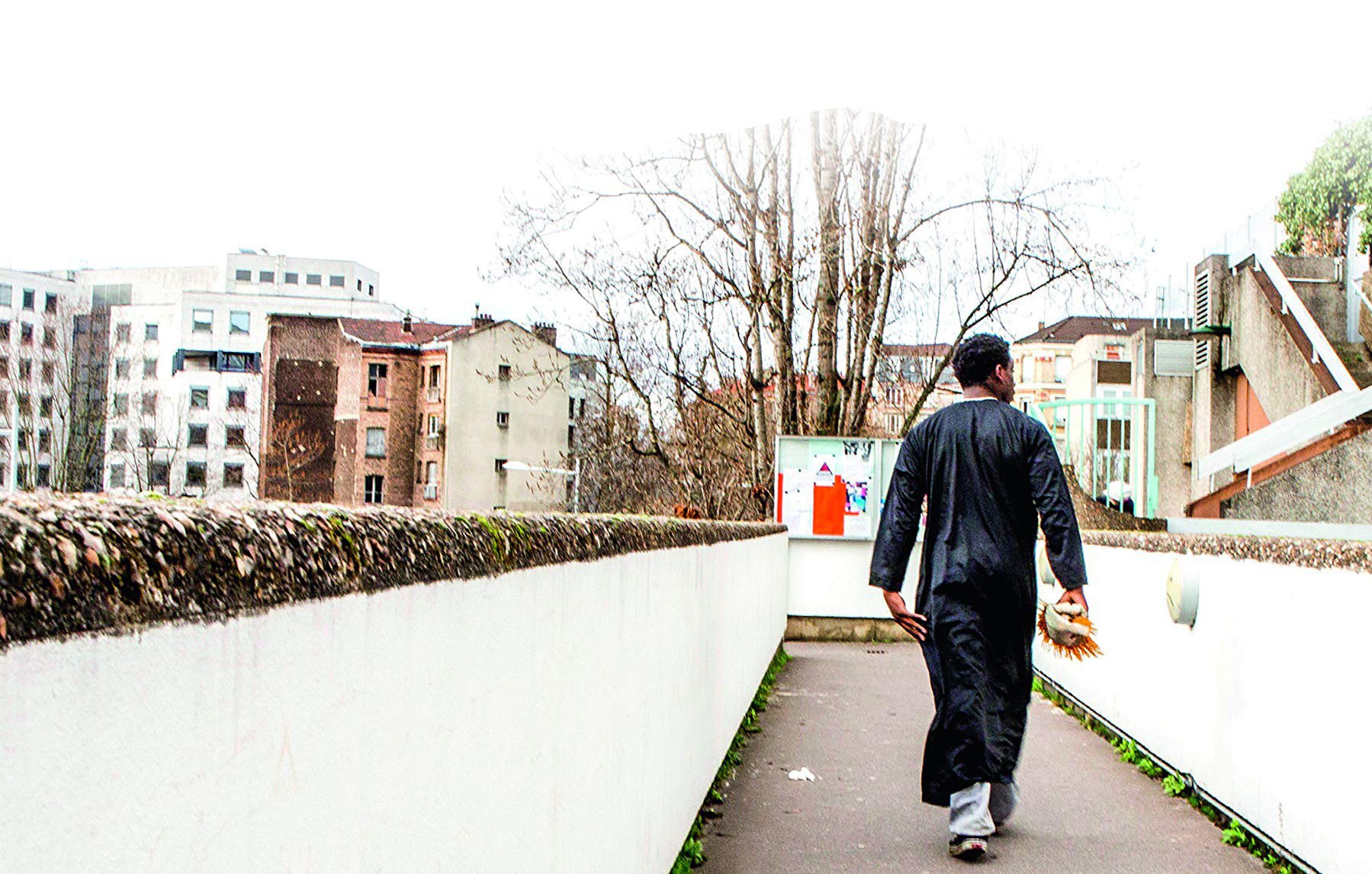 L'image montre un homme marchant sur un chemin urbain. Il porte une tenue traditionnelle, une sorte de longue robe noire. En arrière-plan, on peut voir des bâtiments modernes et anciens, ainsi que des arbres dépouillés. L'environnement semble calme, avec une lumière diffuse, peut-être un jour nuageux. L'homme avance vers l'horizon, de dos, ce qui donne une impression de solitude et de contemplation.