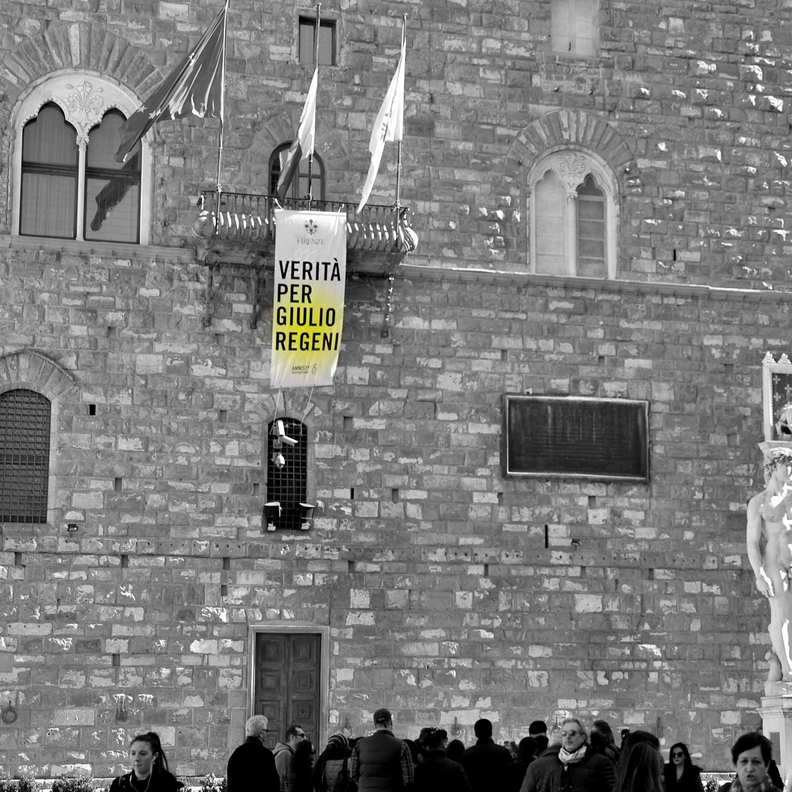 L'image montre un bâtiment historique avec une façade en pierre. Il y a une grande banderole accrochée à la façade, sur laquelle on peut lire "VERITÀ PER GIULIO REGENI", ce qui signifie "Vérité pour Giulio Regeni". En bas, on aperçoit une statue blanche, sûrement une œuvre d'art classique. Plusieurs personnes se déplacent devant le bâtiment, créant une ambiance animée. L'image est en noir et blanc, ce qui accentue le sérieux du message sur la banderole.