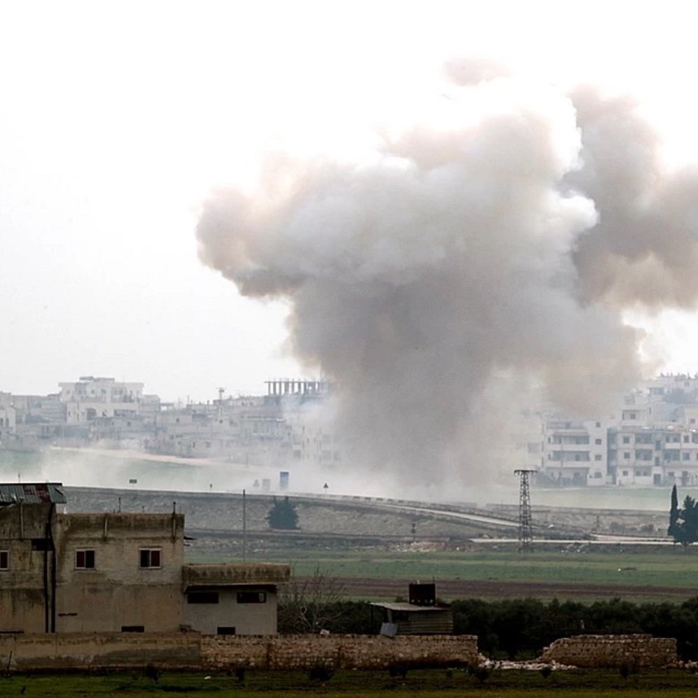 L'image montre un paysage de guerre, avec une explosion qui génère un nuage de fumée sombre s'élevant dans le ciel. Au premier plan, on peut apercevoir des bâtiments endommagés, tandis qu'à l'arrière-plan, une ville est visible, probablement touchée par le conflit. L'atmosphère dégage une impression de destruction et de chaos, illustrant les conséquences d'une situation de conflit armé.