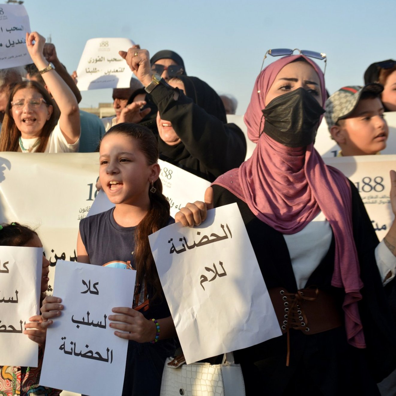 L'image montre un groupe de personnes lors d'une manifestation. Des manifestants, y compris des enfants, tiennent des pancartes avec des inscriptions écrites en arabe. Ils expriment des revendications, visiblement engagés dans une lutte pour une cause qui leur tient à cœur. Certaines personnes portent des masques, et l'ambiance semble déterminée et pleine d'énergie. Le paysage semble ensoleillé, suggérant un rassemblement en plein air.