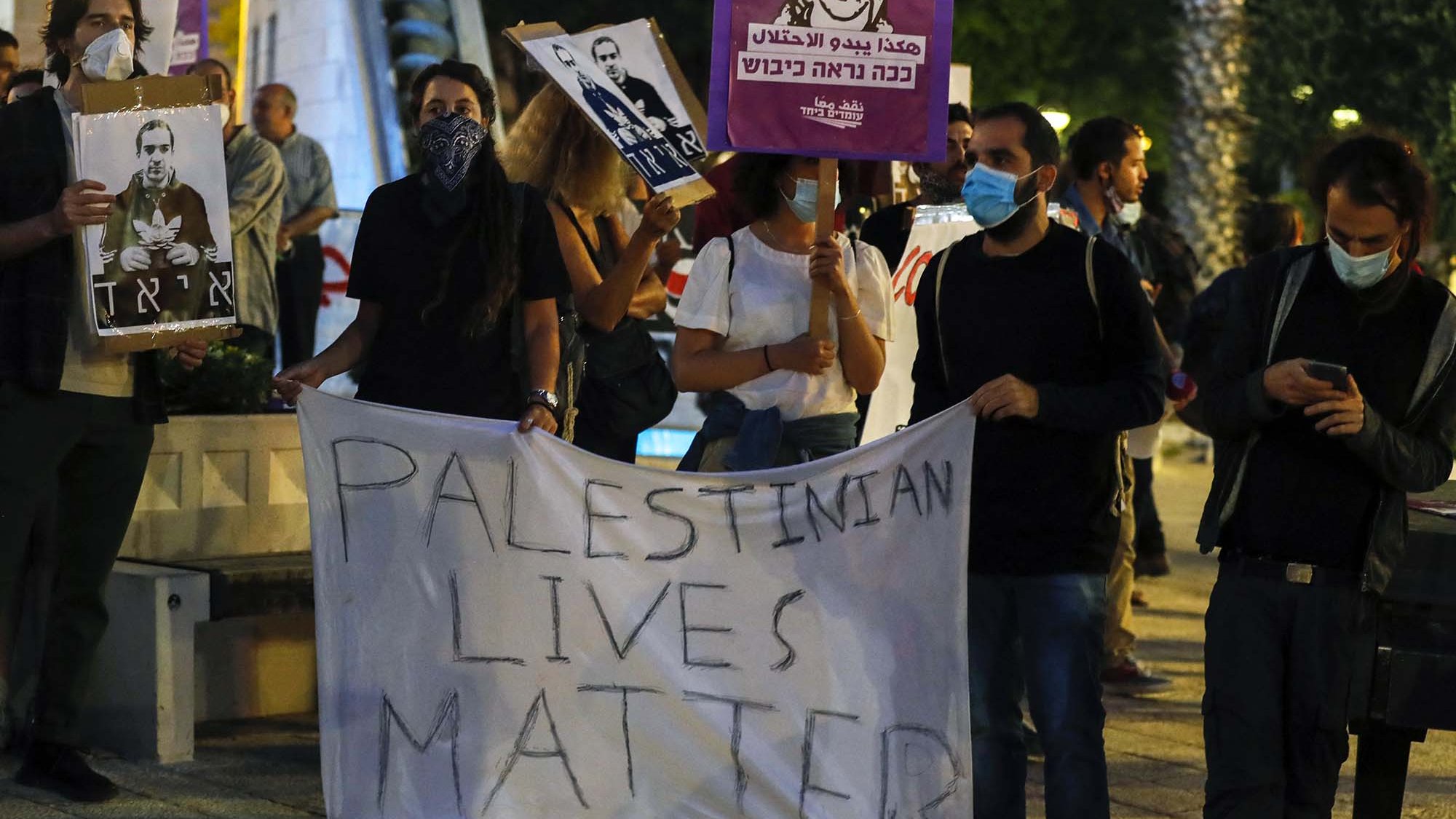 L'image montre un groupe de personnes manifestant. Elles tiennent des panneaux et une grande banderole sur laquelle est écrit "PALESTINIAN LIVES MATTER". Les participants portent des masques, et certains affichent des portraits d'individus. L'ambiance semble être celle d'une manifestation pour les droits des Palestiniens. Le décor urbain, avec des arbres et des lumières en arrière-plan, ajoute à l'atmosphère de l'événement.