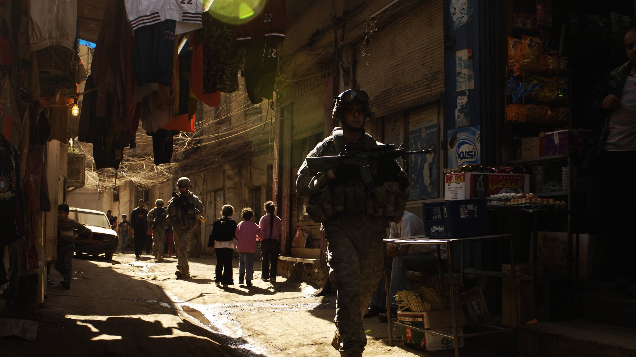 تصویر نشان‌دهنده یک خیابان باریک و شلوغ است که در آن یک سرباز با تجهیزات نظامی در حال حرکت است. نور خورشید از بالای خیابان می‌تابد و سایه‌های طولانی ایجاد کرده است. در دو طرف خیابان، مغازه‌هایی با کالاهای مختلف دیده می‌شوند و چند کودک در حال بازی یا تماشای سربازان هستند. فضای خیابان به طور کلی حس زندگی روزمره و فعالیت‌های شهری را منتقل می‌کند.