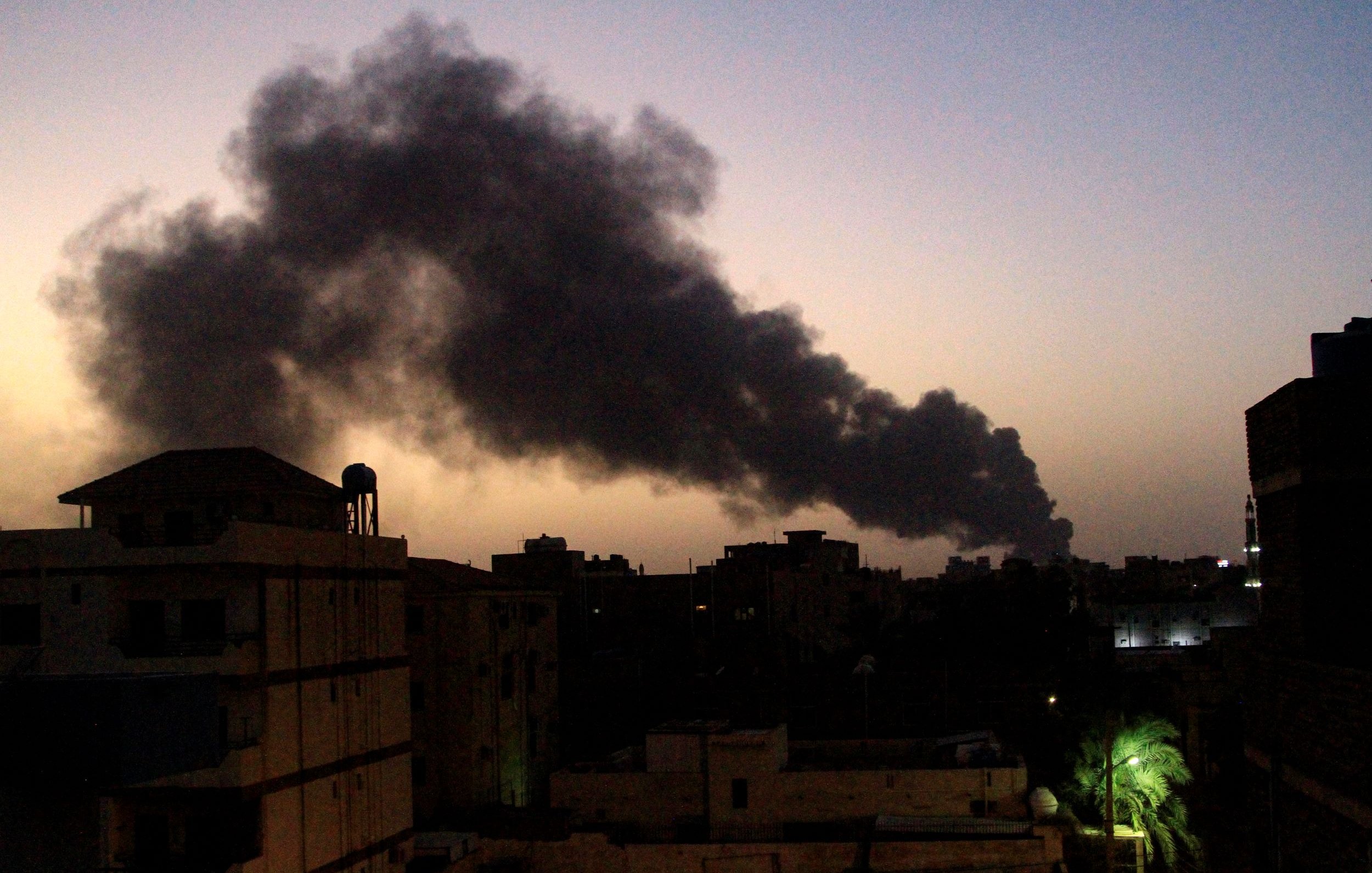 L'immagine mostra un paesaggio urbano al crepuscolo, con un cielo che tende al blu scuro. In lontananza si solleva una colonna di fumo nero, probabilmente proveniente da un incendio o una fonte di combustione. Gli edifici in primo piano sono silhouette scure contro il cielo, e si possono scorgere alcune luci artificiali che brillano tra le strutture. La vegetazione, come le palme, è visibile sul lato destro, aggiungendo un tocco di colore al contesto.