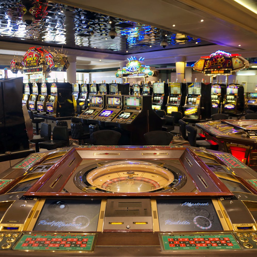 L'image montre l'intérieur d'un casino, avec plusieurs tables de jeux et des machines à sous. Au premier plan, on peut voir une grande table de roulette, entourée de plusieurs sièges. À l'arrière-plan, on distingue de nombreuses machines à sous illuminées, créant une atmosphère animée et colorée. Le plafond est décoré, ajoutant à l'élégance de l'espace. L'ensemble dégage une ambiance de divertissement et de luxe.