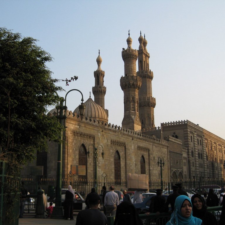 L'image montre un vaste bâtiment architectural au style islamique, probablement une mosquée, avec des minarets majestueux s'élevant vers le ciel. On peut voir des personnes en tenue traditionnelle, certaines portant le hijab. En arrière-plan, la lumière du soleil est douce, créant une atmosphère paisible. Les détails architecturaux de la façade en pierre ajoutent une touche historique à la scène. Des arbres et des lampadaires embellissent également l'environnement.