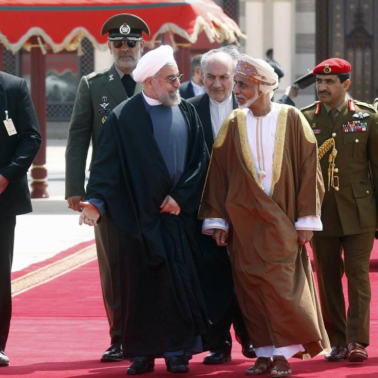The image features a group of men walking together on a red carpet, likely in a formal or diplomatic setting. In the foreground, two men are engaged in conversation, one wearing a black suit and the other in traditional attire. They appear to be leaders or dignitaries. Behind them, there are several men in military uniforms and suits, possibly security personnel or officials, accompanying the two main figures. The background suggests an ornate architectural setting, enhancing the official nature of the gathering.