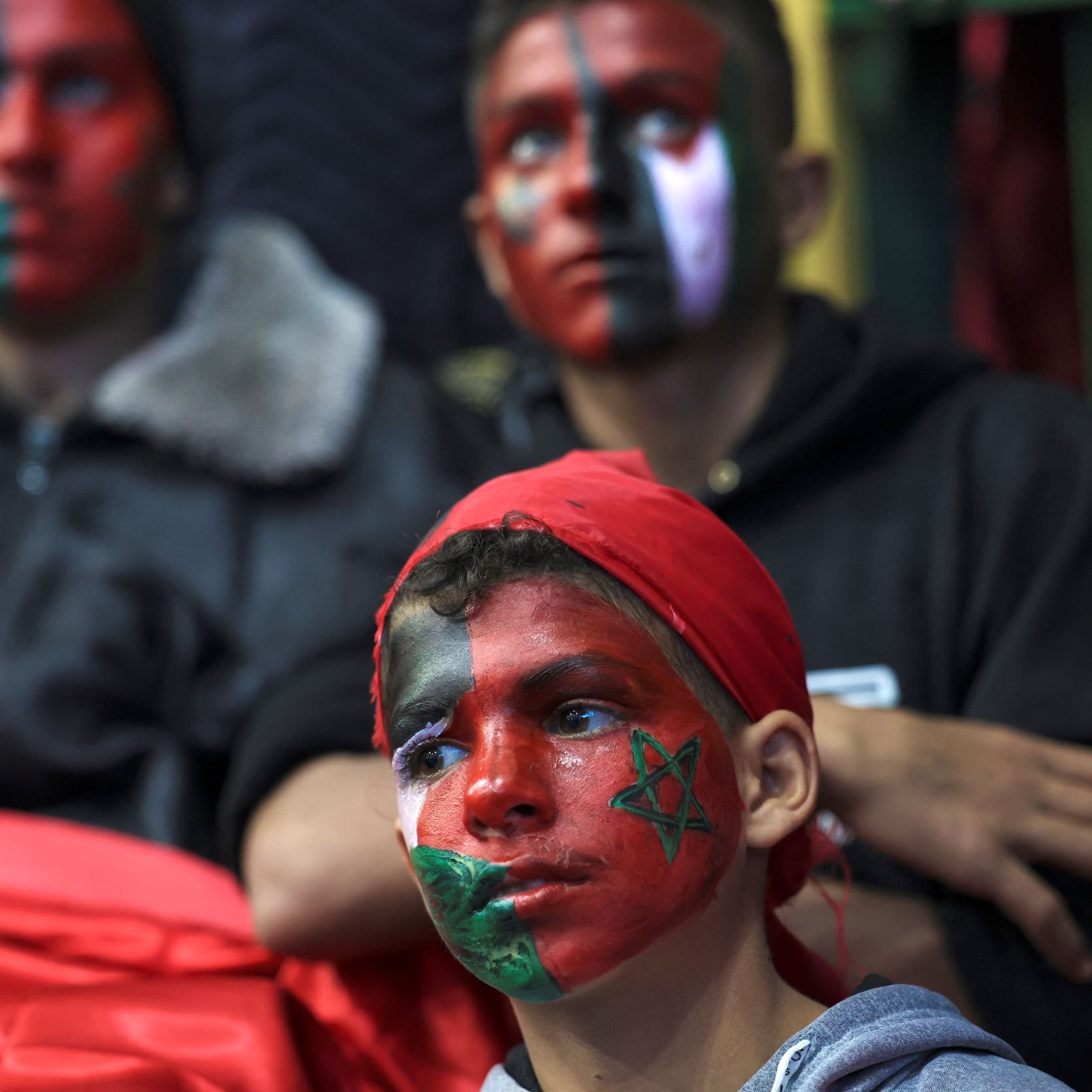 L'image montre un groupe de jeunes, dont un enfant au premier plan, qui ont le visage peint avec des motifs aux couleurs du drapeau marocain. L'enfant porte un bandana rouge et des dessins représentant le drapeau marocain et d'autres motifs sur son visage. En arrière-plan, deux autres personnes affichent également des visages peints, créant une atmosphère festive et engagée. L'ambiance semble liée à un événement de soutien ou à une célébration.