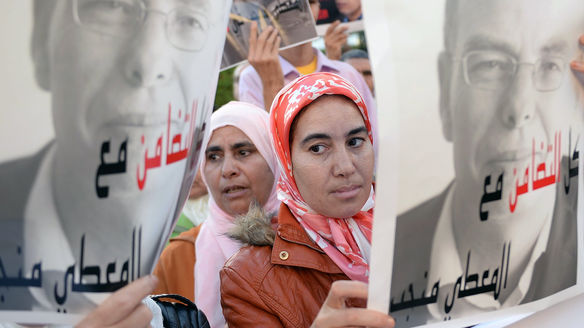 L'image présente des manifestants tenant des pancartes. Au premier plan, une femme avec un foulard rose regarde l'objectif, tandis qu'une autre femme, en arrière-plan, semble également attentive. Les pancartes affichent des portraits en noir et blanc d'un homme, accompagnés de slogans en arabe. L'ambiance suggère un rassemblement ou une manifestation, probablement pour défendre une cause ou exprimer une opinion.