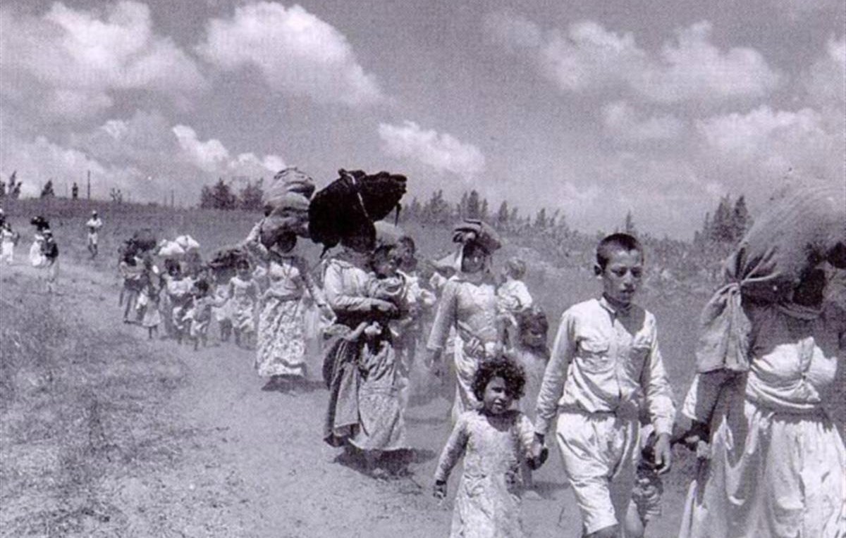 L'image montre un groupe de personnes marchant sur un chemin. On y voit des hommes, des femmes et des enfants, portant des charges sur leur tête ou dans leurs bras. Les adultes semblent laborieux, tandis que les enfants marchent à leurs côtés. Le paysage est rural, avec un ciel nuageux en arrière-plan, et l'atmosphère générale évoque une situation de déplacement ou d'exode. Les vêtements des personnes sont simples et évoquent une époque passée.