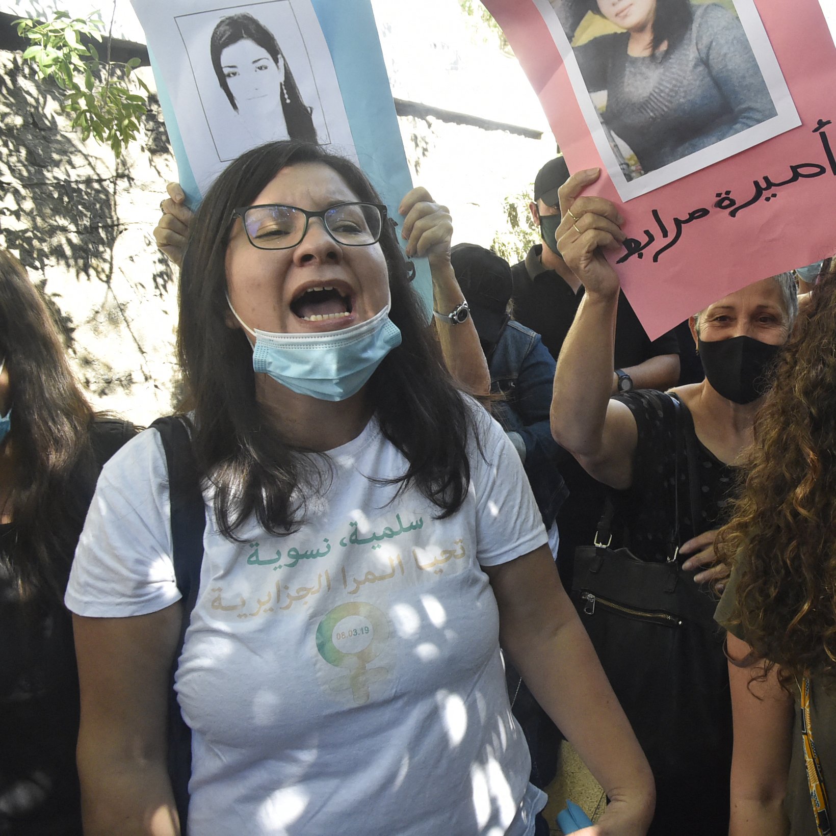 در این تصویر، گروهی از زنان و مردان در حال برگزاری یک تظاهرات هستند. آن‌ها پلاکاردهایی در دست دارند که تصاویری از افراد مختلف بر روی آن‌ها چاپ شده است. برخی از شرکت‌کنندگان به شدت فریاد می‌زنند و احساسات خود را ابراز می‌کنند. اکثر افراد ماسک به چهره دارند، که به خاطر وضعیت بهداشتی ممکن است باشد. فضای اطراف شلوغ به نظر می‌رسد و به وضوح نشان‌دهنده یک حرکت اجتماعی است.