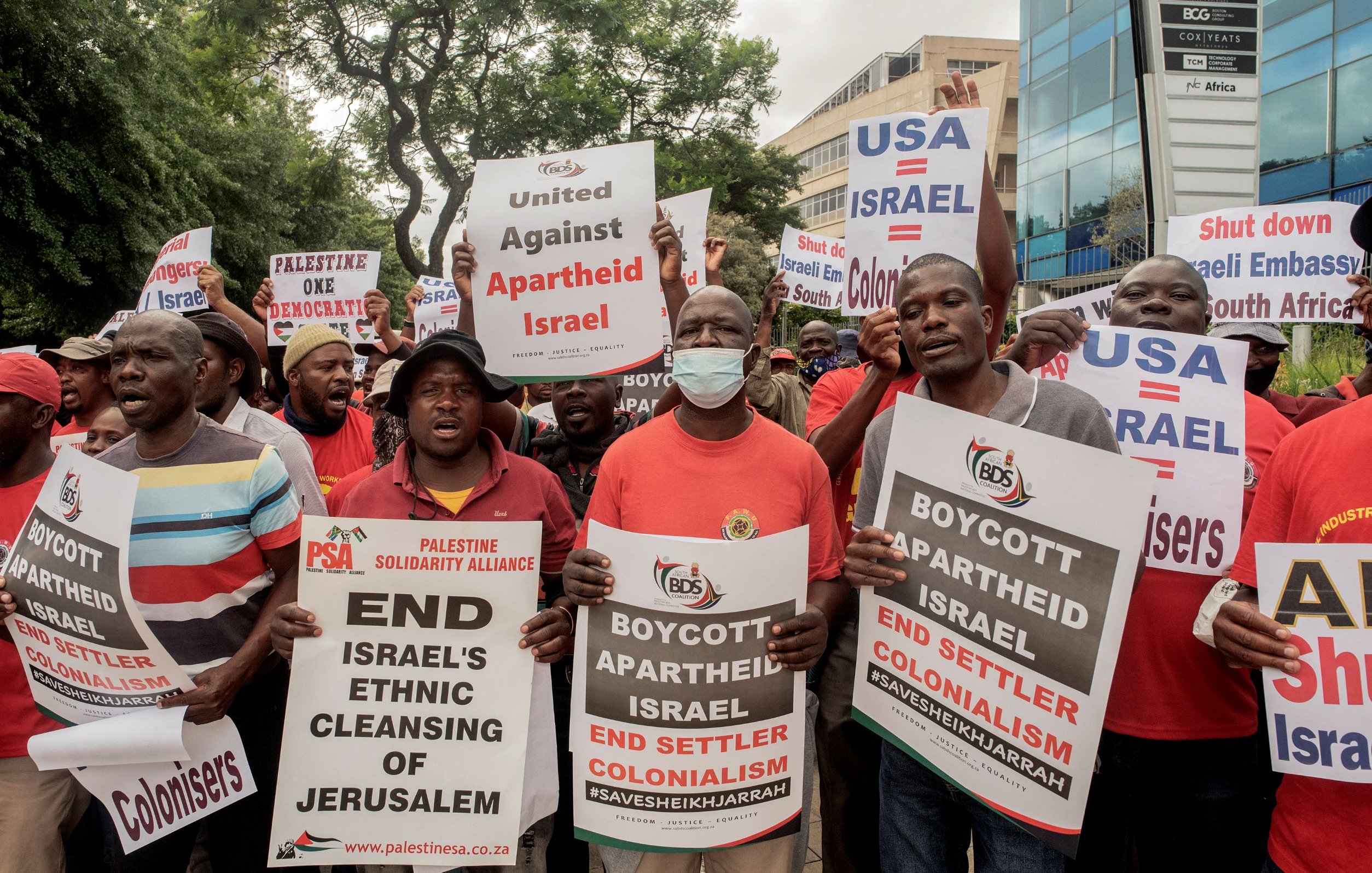 L'image montre une foule de manifestants tenant des pancartes et exprimant leur opposition à la politique israélienne, en particulier en ce qui concerne le traitement des Palestiniens. Les slogans sur les pancartes incluent des appels à mettre fin à l'apartheid et au colonialisme en Israël, ainsi qu'à boycotter certaines actions liées à ce pays. Les manifestants semblent très engagés dans leur cause, et l'atmosphère est dynamique et déterminée.