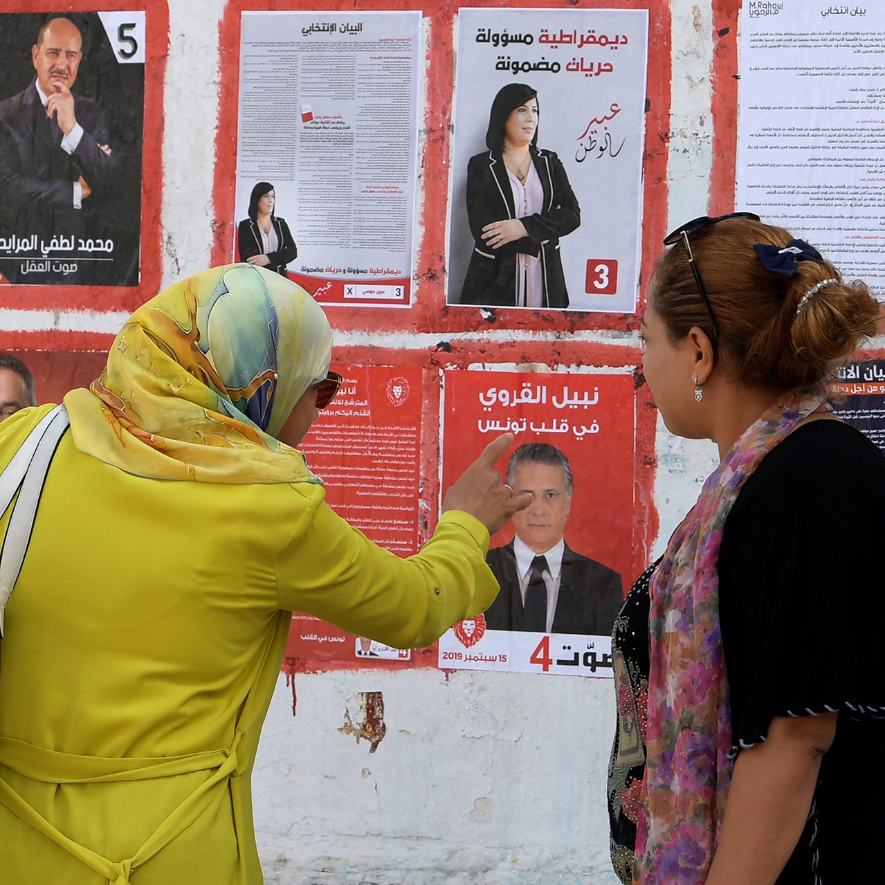 L'image montre un mur recouvert d'affiches électorales. Deux femmes, dont l'une porte un foulard, discutent et pointent une des affiches. Les affiches présentent des candidats avec leurs photos et informations pertinentes, probablement dans le cadre d'une élection. L'atmosphère semble être celle d'une campagne électorale, avec un intérêt évident pour les candidats et leurs programmes.