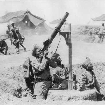 L'image en noir et blanc montre un groupe de soldats en position de tir. Un soldat est en train d'opérer un mortier, levé vers le ciel, tandis que deux autres soldats se trouvent à proximité, probablement prêts à aider ou à surveiller la situation. En arrière-plan, on peut voir des tentes militaires, indiquant qu'ils sont dans un camp. D'autres soldats semblent en mouvement, suggérant une activité intense ou une opération en cours. L'atmosphère générale de l'image évoque un contexte militaire et historique.