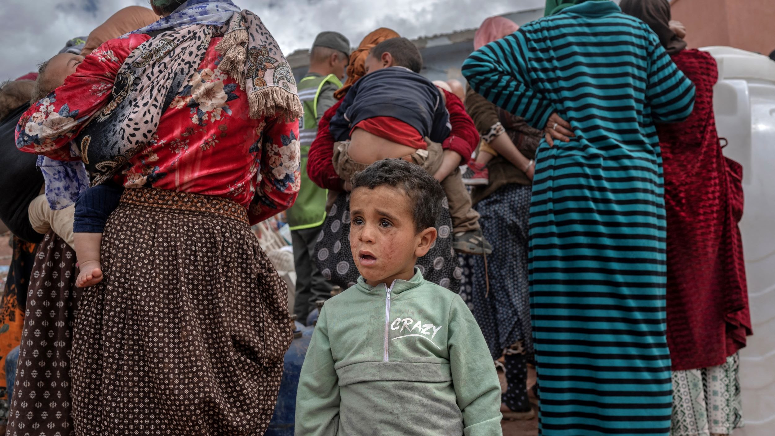 La imagen muestra a un grupo de personas de espaldas, predominantemente mujeres, que parecen estar en un ambiente rural. La ropa que llevan es variada, con patrones y colores diversos. En el primer plano, un niño pequeño aparece al frente, mirando hacia la cámara con una expresión que podría reflejar confusión o preocupación. El fondo sugiere un cielo nublado y la escena evoca una sensación de comunidad y un posible evento o reunión.
