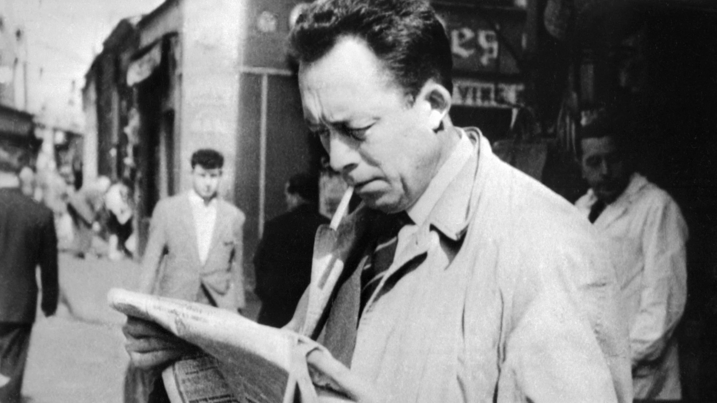 L'immagine ritrae un uomo in primo piano che legge un giornale, con una sigaretta tra le labbra. Indossa un soprabito chiaro e sembra assorto nella lettura. Sullo sfondo, si possono vedere altre persone che camminano per strada, in un contesto urbano. L'atmosfera è retro e richiama un'epoca passata, con un'illuminazione naturale e dettagli architettonici tipici di quel periodo.