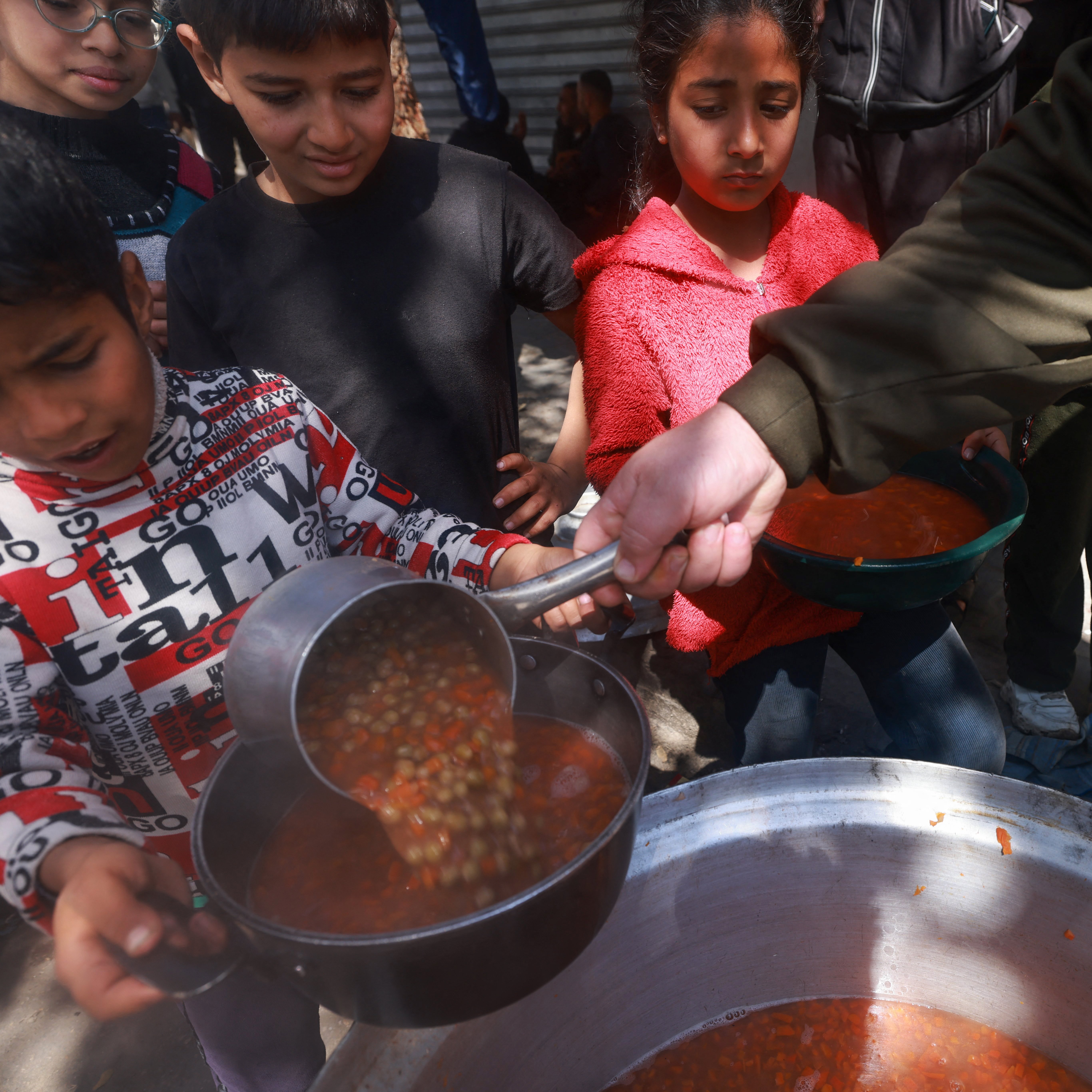 L'image montre des enfants qui se rassemblent autour d'une grande marmite. Un enfant, tenant une louche, verse une soupe dans un bol. D'autres enfants, attentifs, attendent leur tour pour recevoir leur repas. L'atmosphère semble conviviale, et l'environnement est extérieur, probablement dans une aire où des repas sont servis. Les enfants portent des vêtements variés, et certains semblent impatients et enthousiastes.