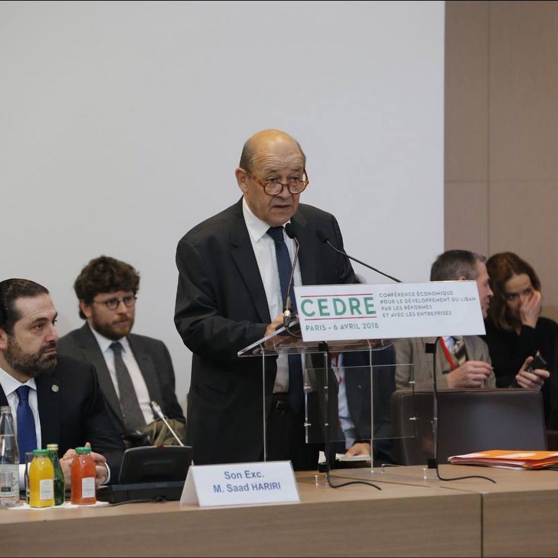 L'image montre un homme âgé, probablement un orateur, se tenant derrière un podium avec un panneau portant le mot "CEDRE" et la date de l'événement. Il semble prendre la parole lors d'une conférence. Autour de lui, plusieurs autres personnes sont assises, attentives, et il y a des bouteilles d'eau et des boissons sur la table. L'ambiance semble formelle et professionnelle, suggérant un événement important.